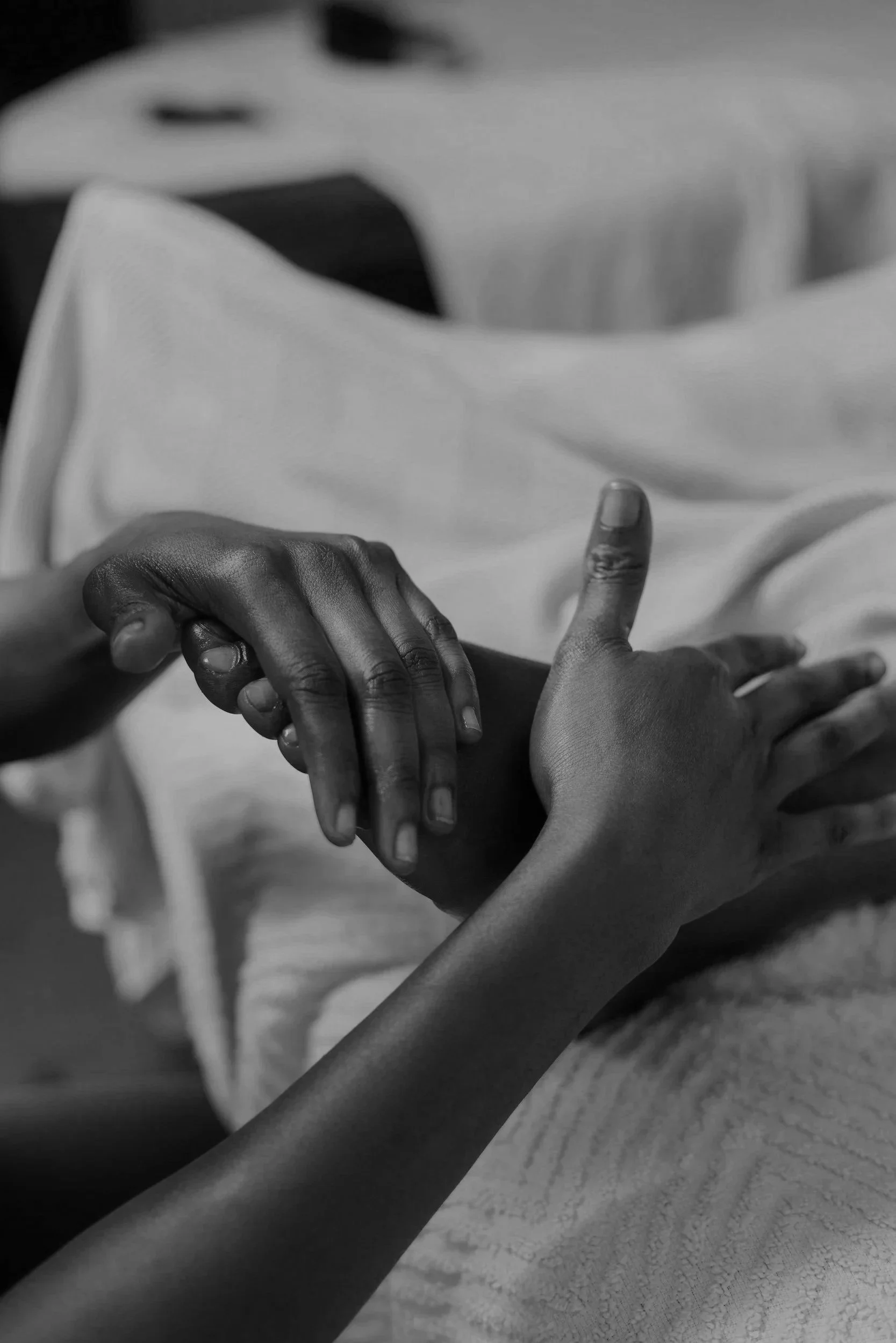 Close-up black-and-white photo of two hands holding each other, one with darker skin and the other with lighter skin, on a table or surface.