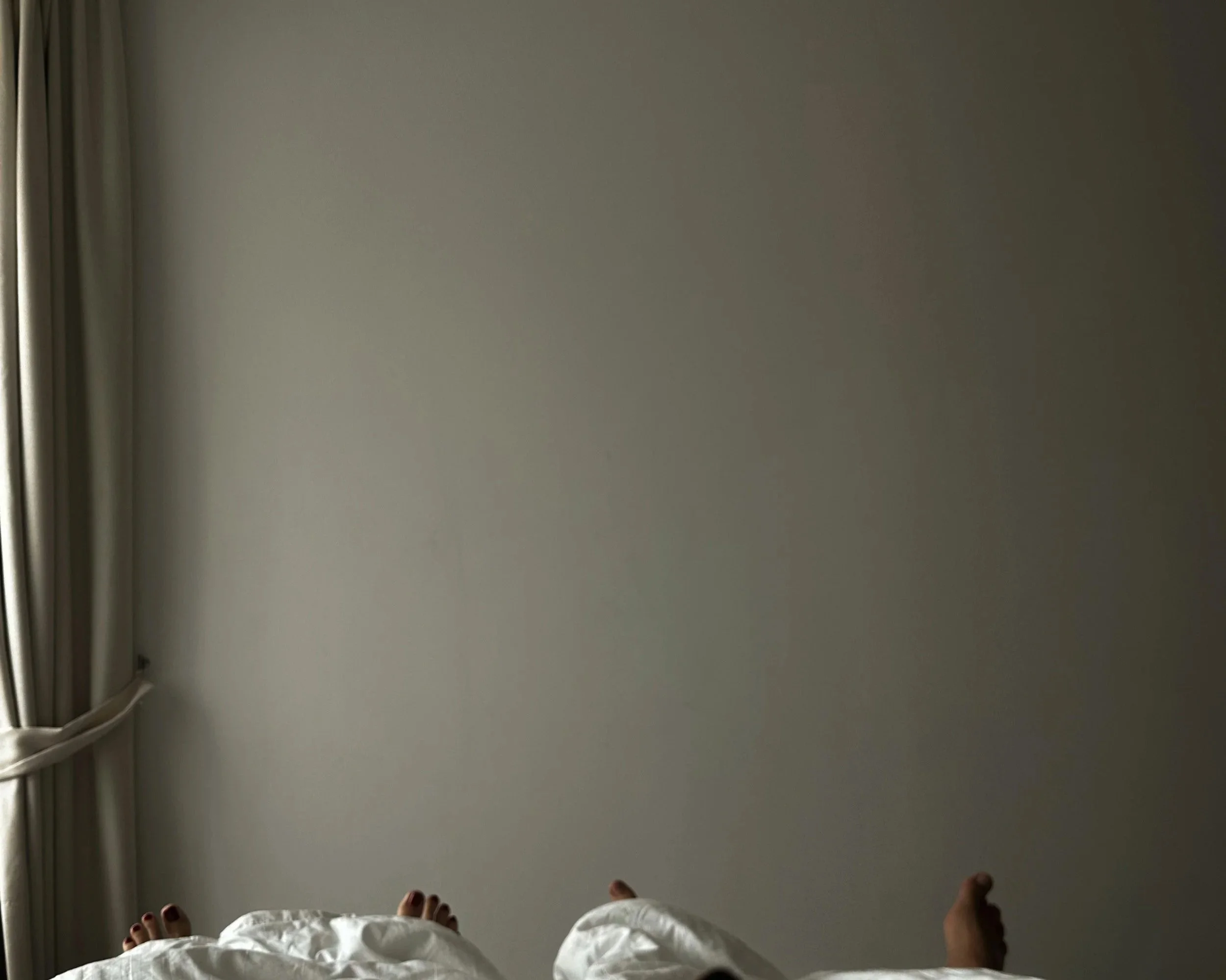Photo of a person standing on a white bed with a gray wall in the background, view from their perspective, toes visible, and part of a curtain on the left.