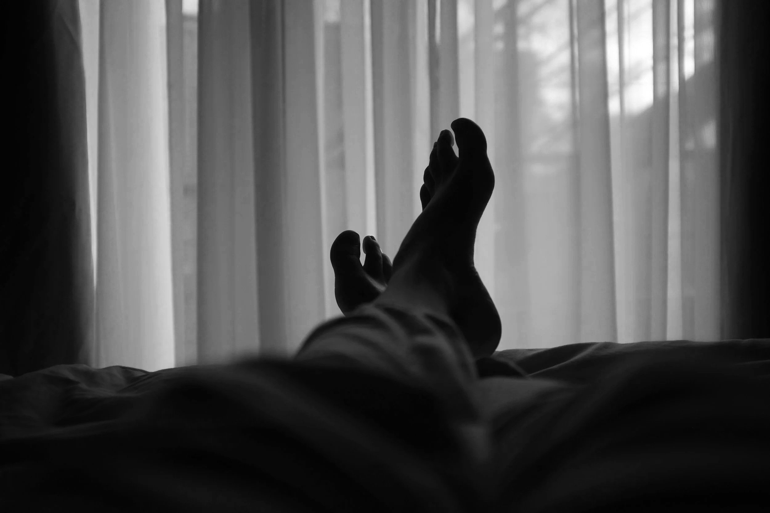 Black and white photo showing a person reaching out their hand towards a window with curtains, with trees visible outside.