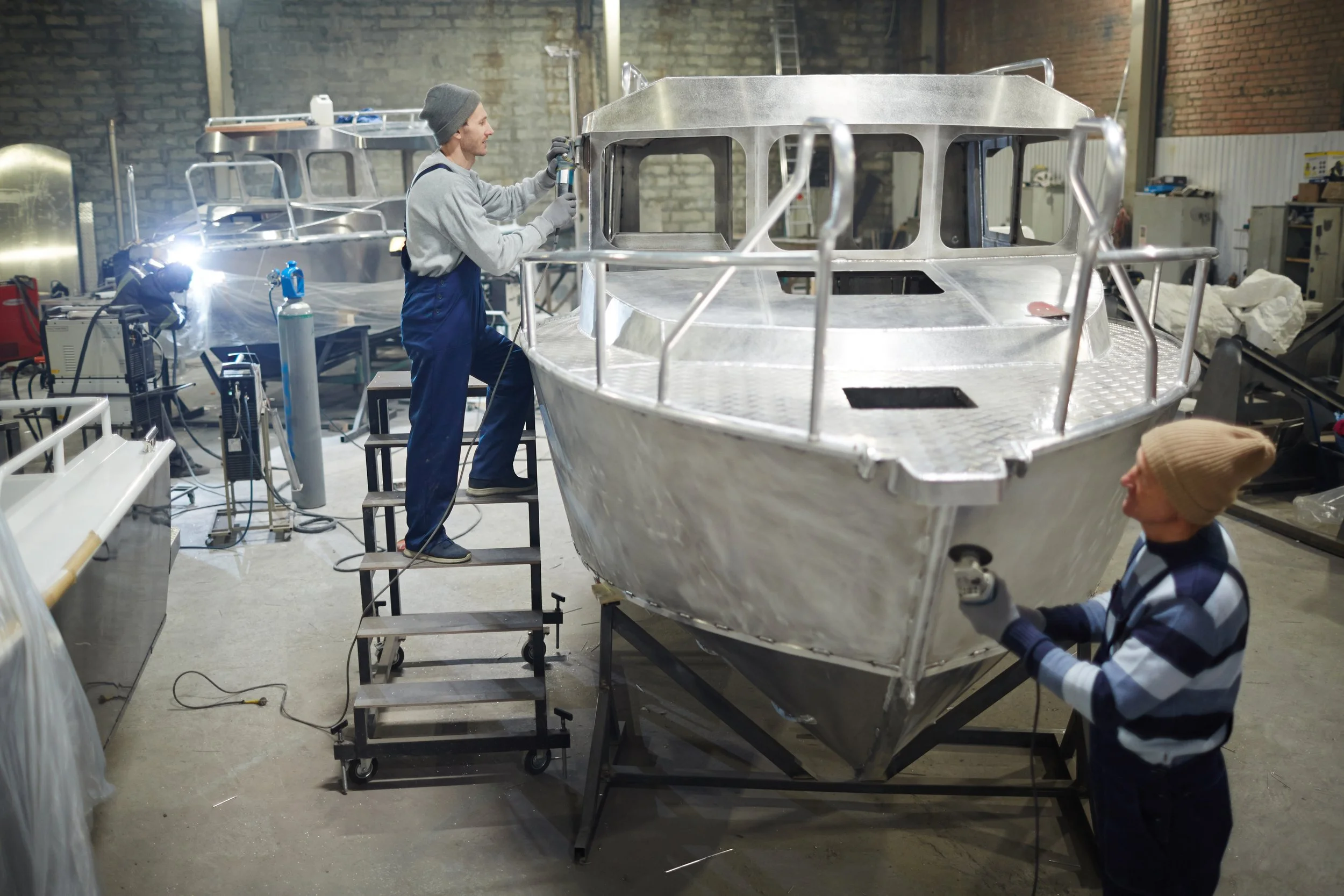 A person in work overalls and a beanie, welding on a metal boat in an industrial workshop.
