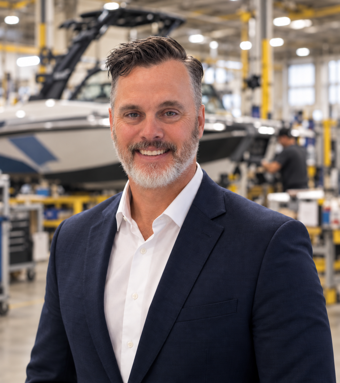 A middle-aged man with gray hair and a beard, wearing a navy blue suit and white shirt, smiling in a manufacturing or assembly plant with a boat or yacht in the background.