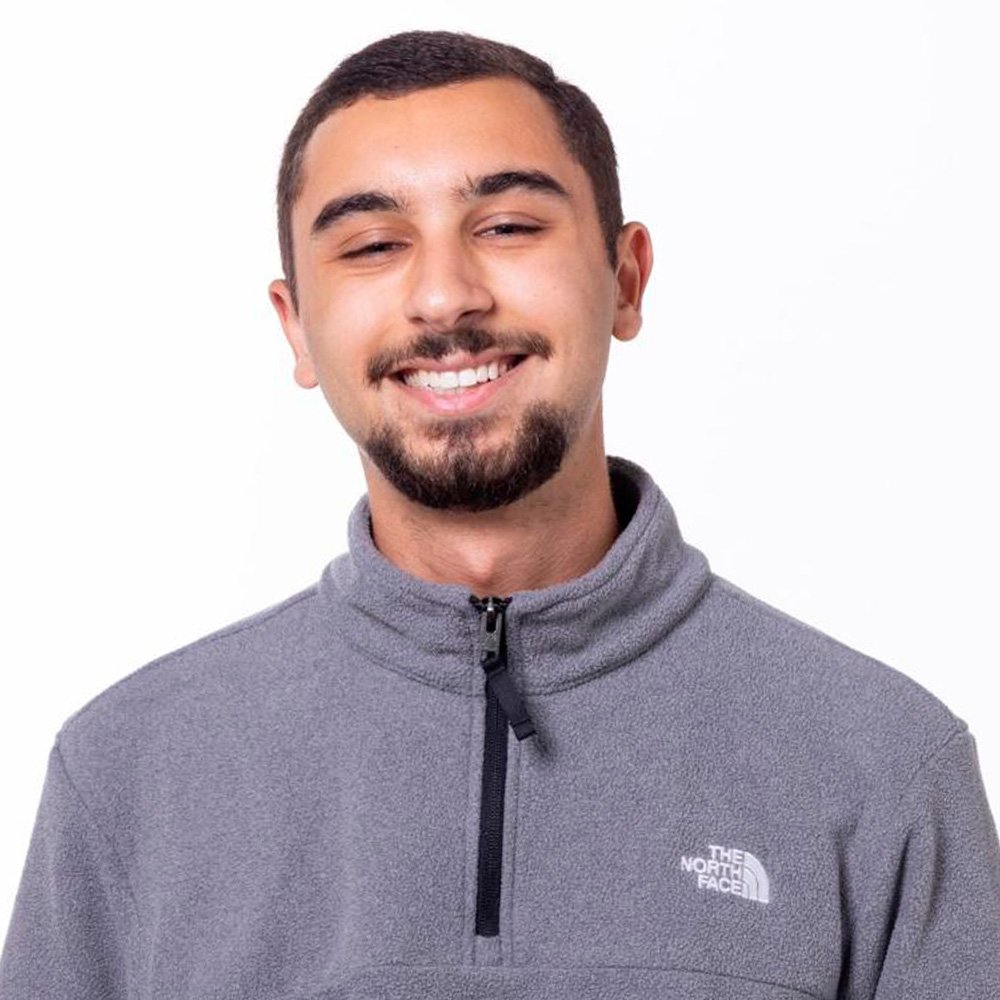 Smiling young man with short dark hair and a beard, wearing a gray North Face quarter-zip fleece jacket, standing against a white background.