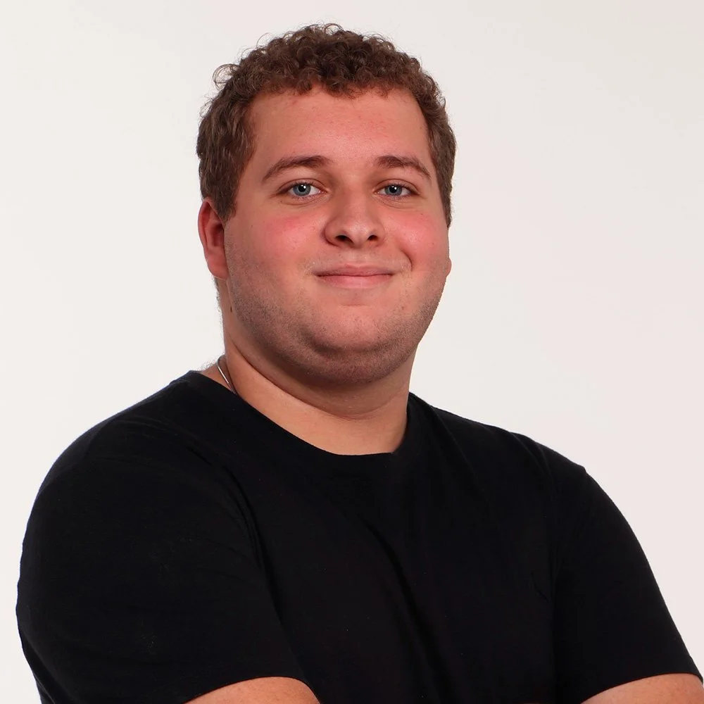 A young man with short, curly brown hair and blue eyes is smiling and looking at the camera. He is wearing a plain black T-shirt and standing against a white background.