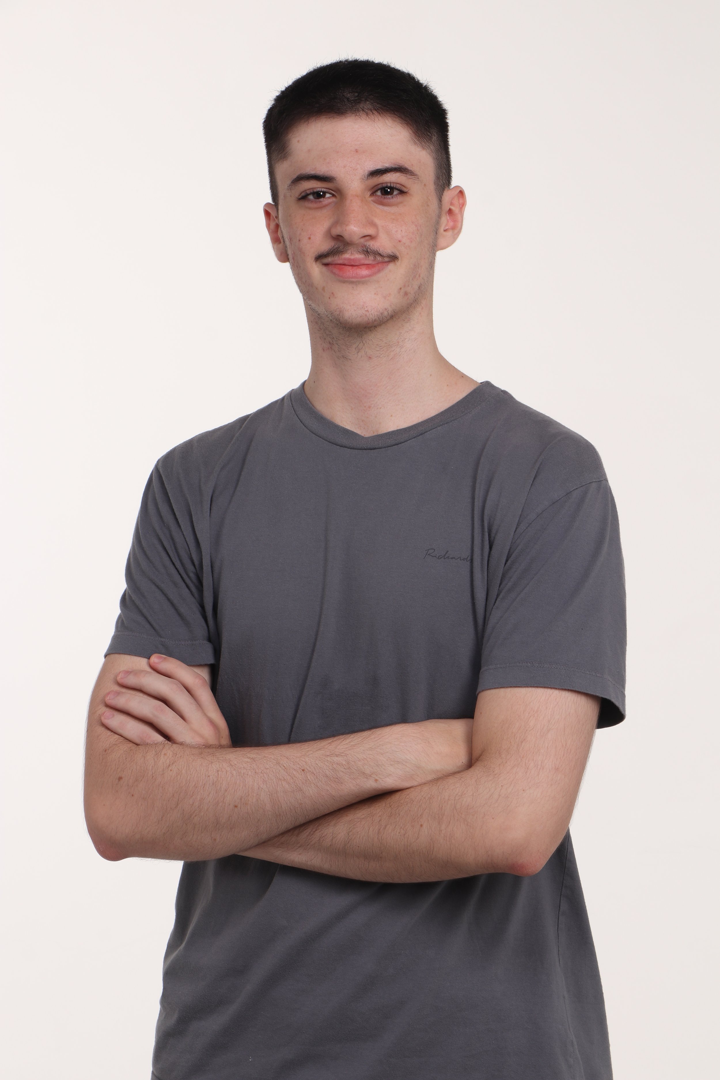 A young man with short dark hair, light skin, and some facial acne, standing with arms crossed, wearing a gray t-shirt, posing against a plain off-white background.