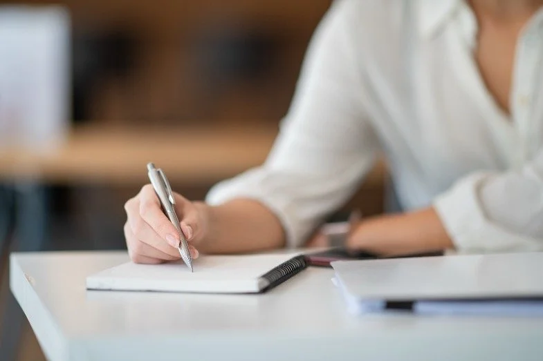 A person wearing a white shirt is writing in a notebook with a silver pen, with other papers on a white table.
