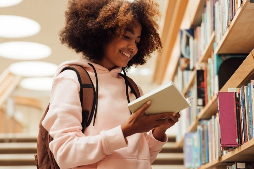 A young woman with curly hair smiling while reading a book in a library aisle.