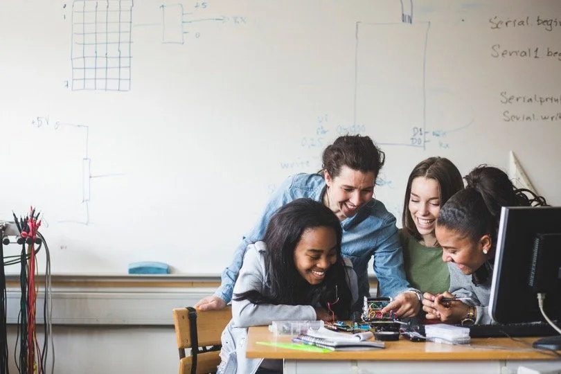 Four young women gathered around a table, smiling and working together on electronics projects in a classroom with a whiteboard and computer.