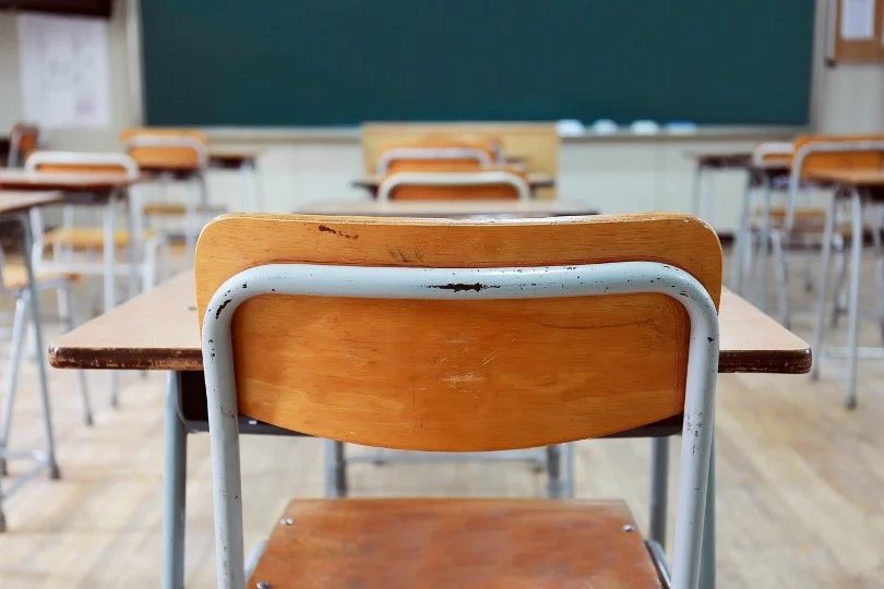 Empty classroom with wooden desks and chairs, and a chalkboard in the background.
