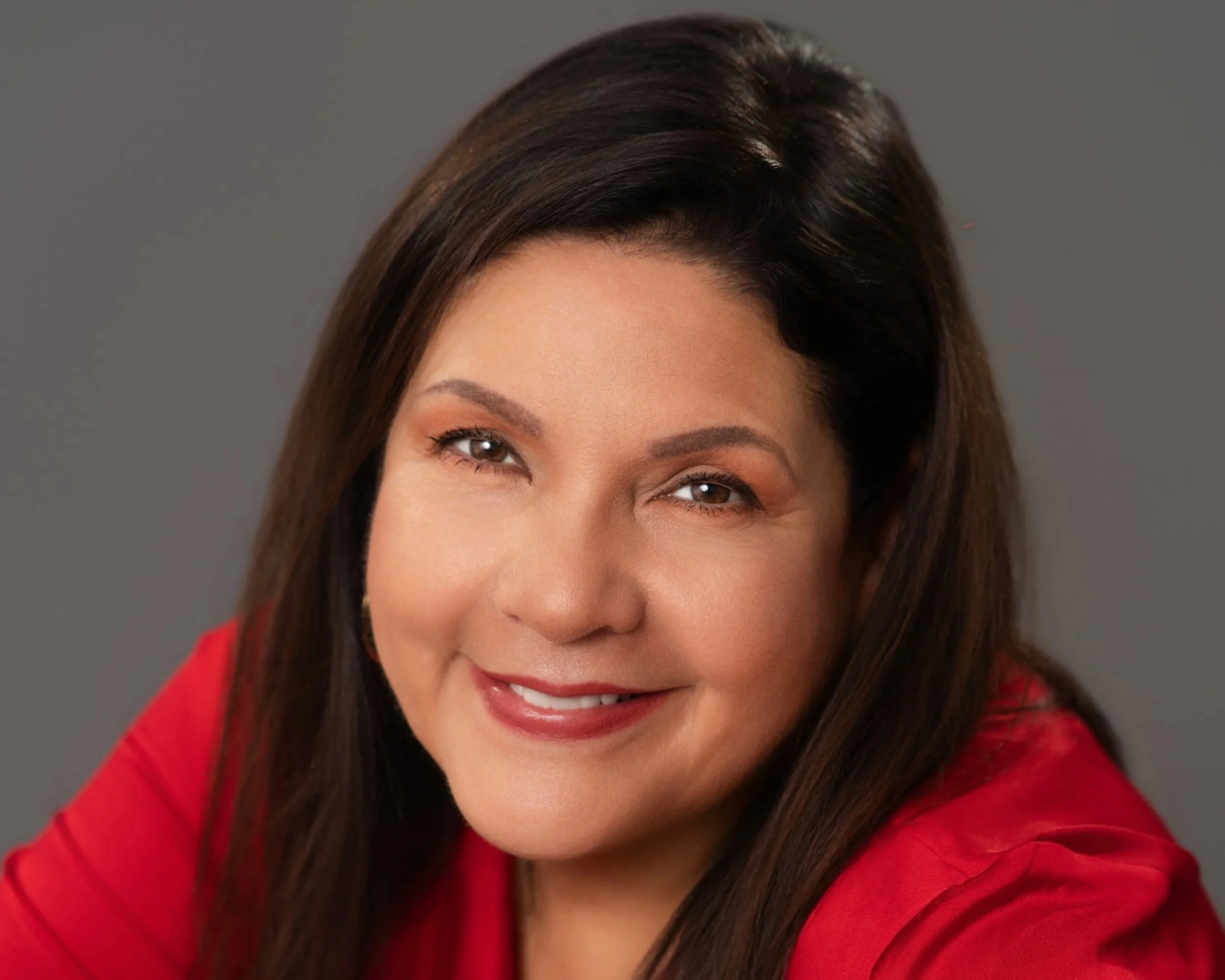 Close-up of a smiling woman with long brown hair, wearing a red top, against a gray background.