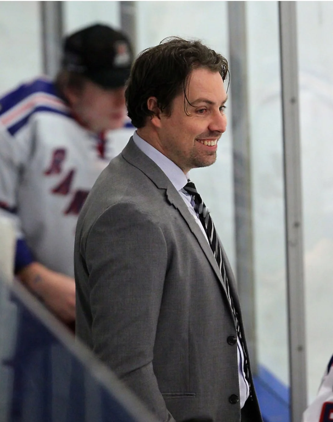 A man in a gray suit with a striped tie smiling on the ice rink, with hockey players in the background.