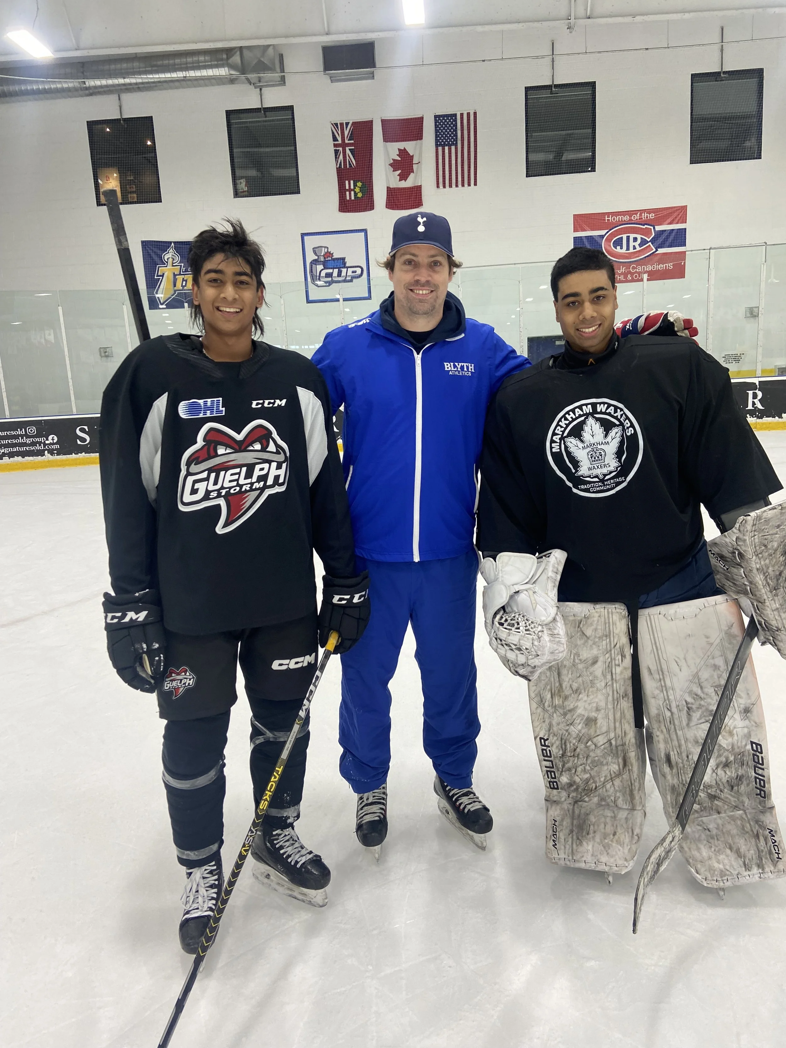 Three men standing on an ice hockey rink together, smiling at the camera. The man in the middle is wearing a blue jacket and cap, while the two men on either side are wearing black hockey jerseys with logos. The background features an ice rink with Canadian and American flags hanging on the wall.