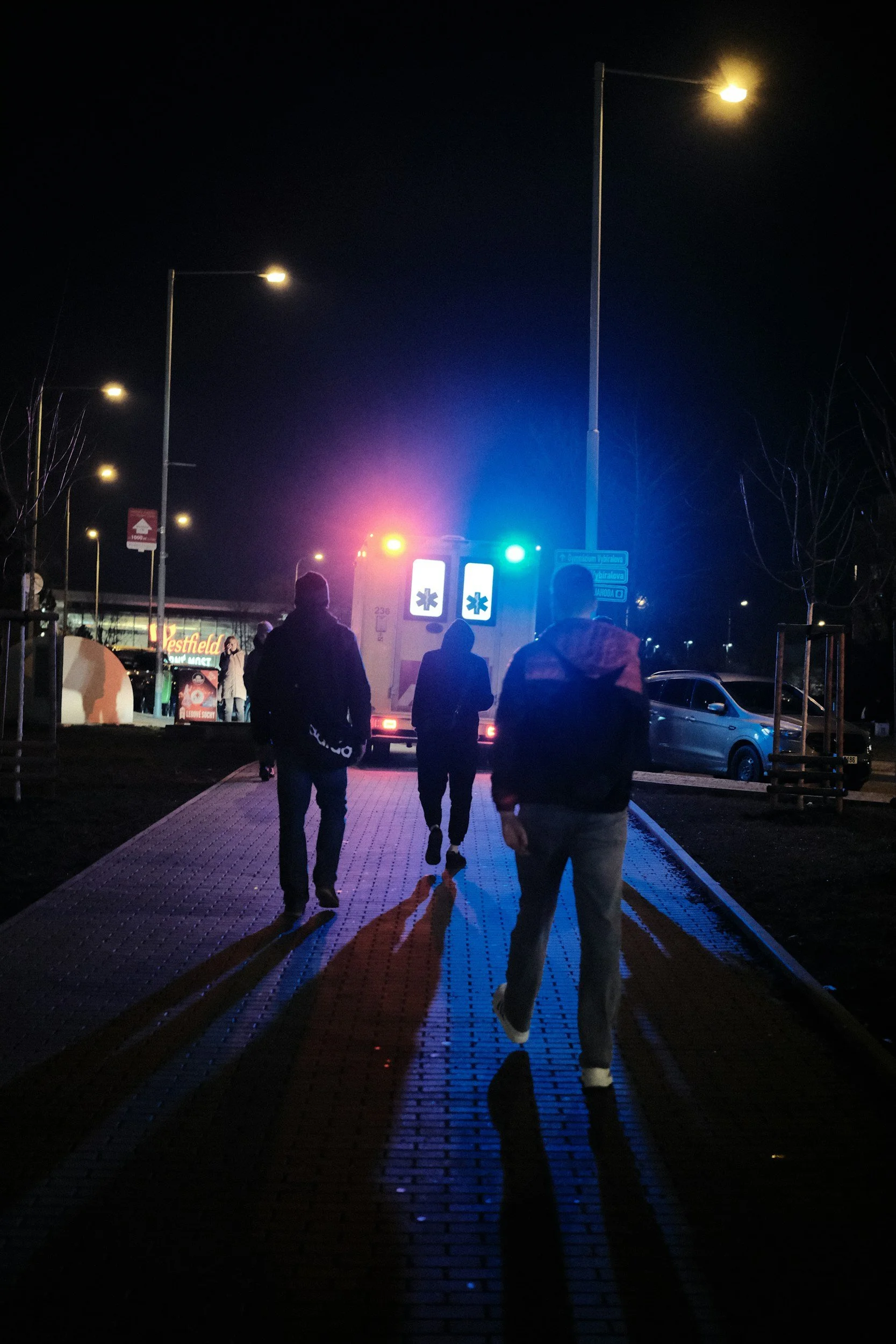 Nighttime scene of people walking on a lit sidewalk with an ambulance nearby and police lights flashing in the background.