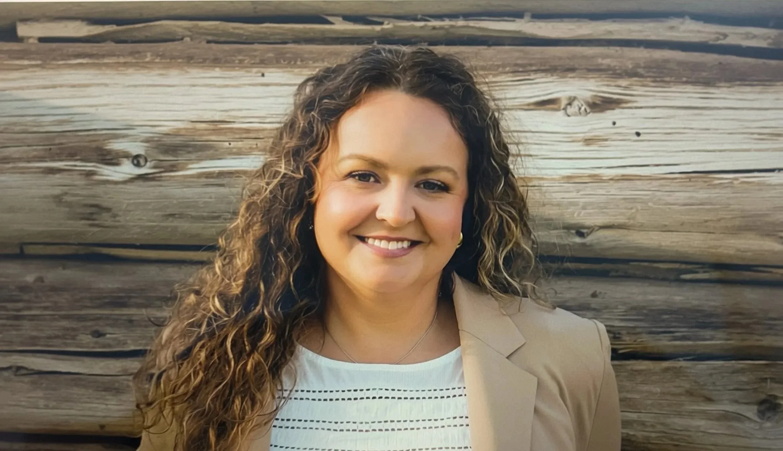 A woman with long, curly hair smiling in front of a wooden wall, wearing a light-colored blazer and a white top.