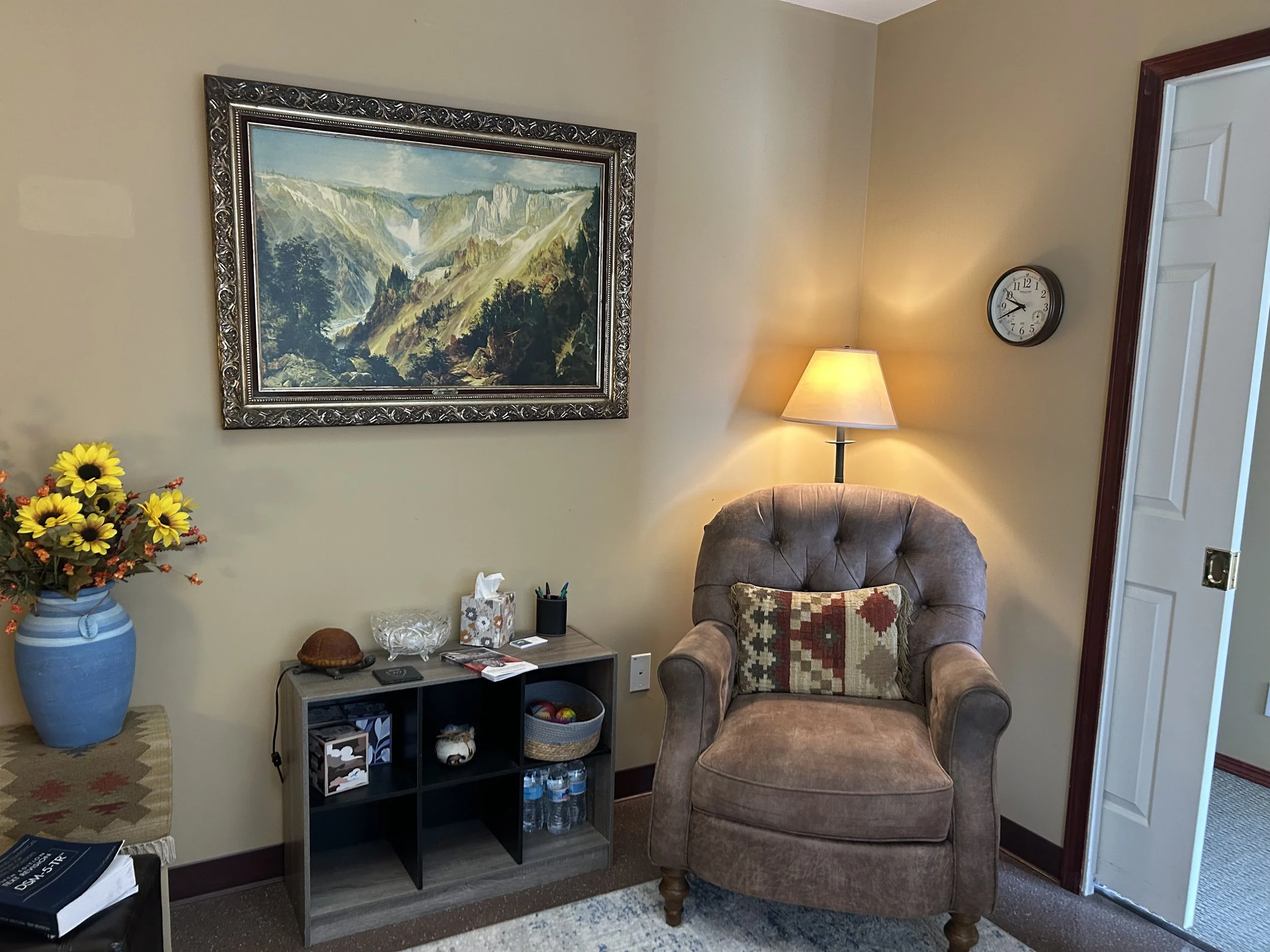 Living room corner with a framed mountain landscape painting, a table lamp emitting warm light, a plush armchair with a decorative pillow, a blue vase with flowers, and a small shelf holding water bottles and books.