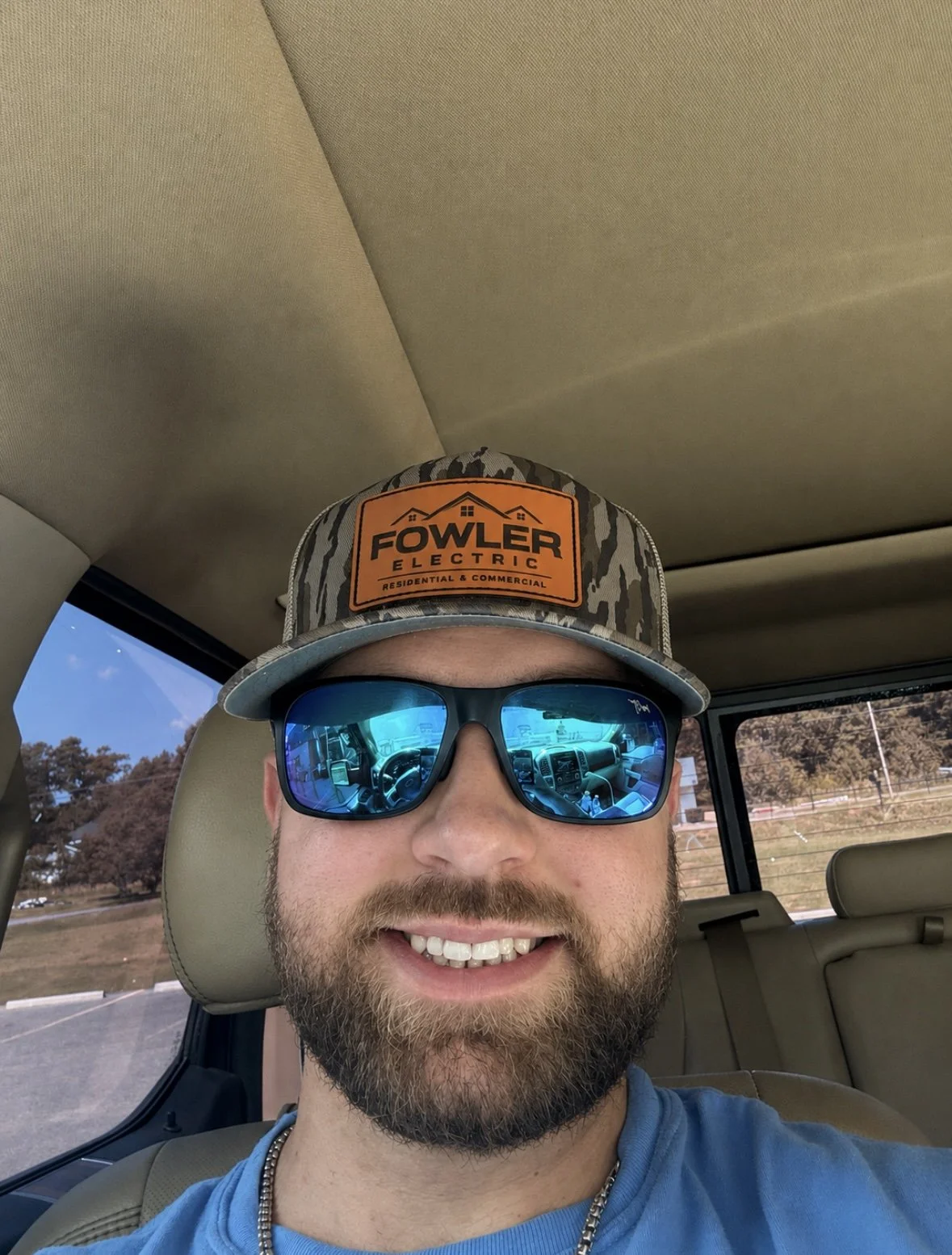 A man with a beard and wearing sunglasses is smiling in a vehicle. He is wearing a camo baseball cap with an orange patch that reads 'Fowler Electric Residential & Commercial.' The vehicle's interior and a sunny outdoor scene with trees and a fence are visible.