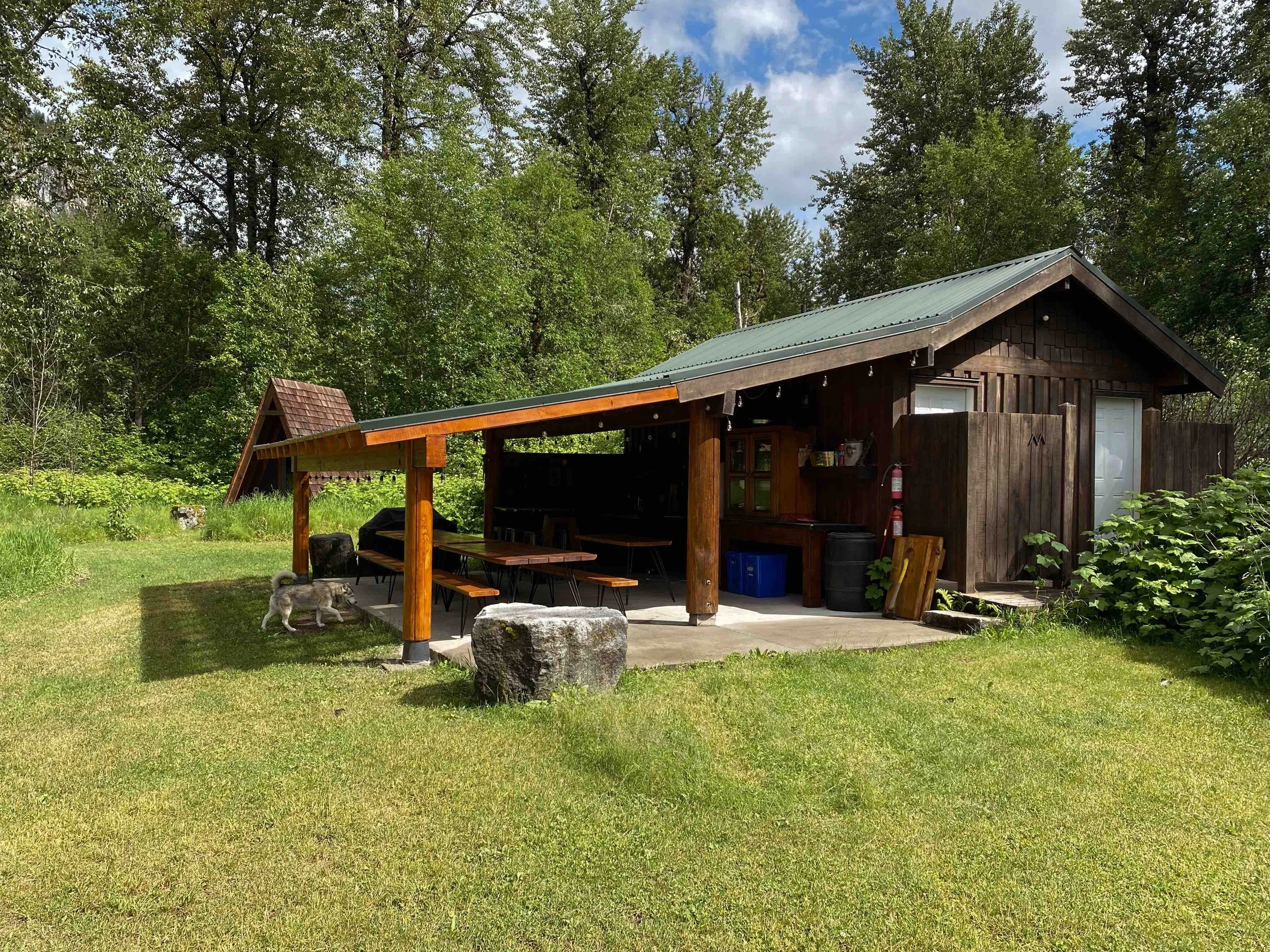A small wooden house or cabin with a sloped metal roof, situated in a lush green yard with trees in the background. There is a covered area with a wooden picnic table and benches, a fire extinguisher, and a Siberian Husky dog in the yard.