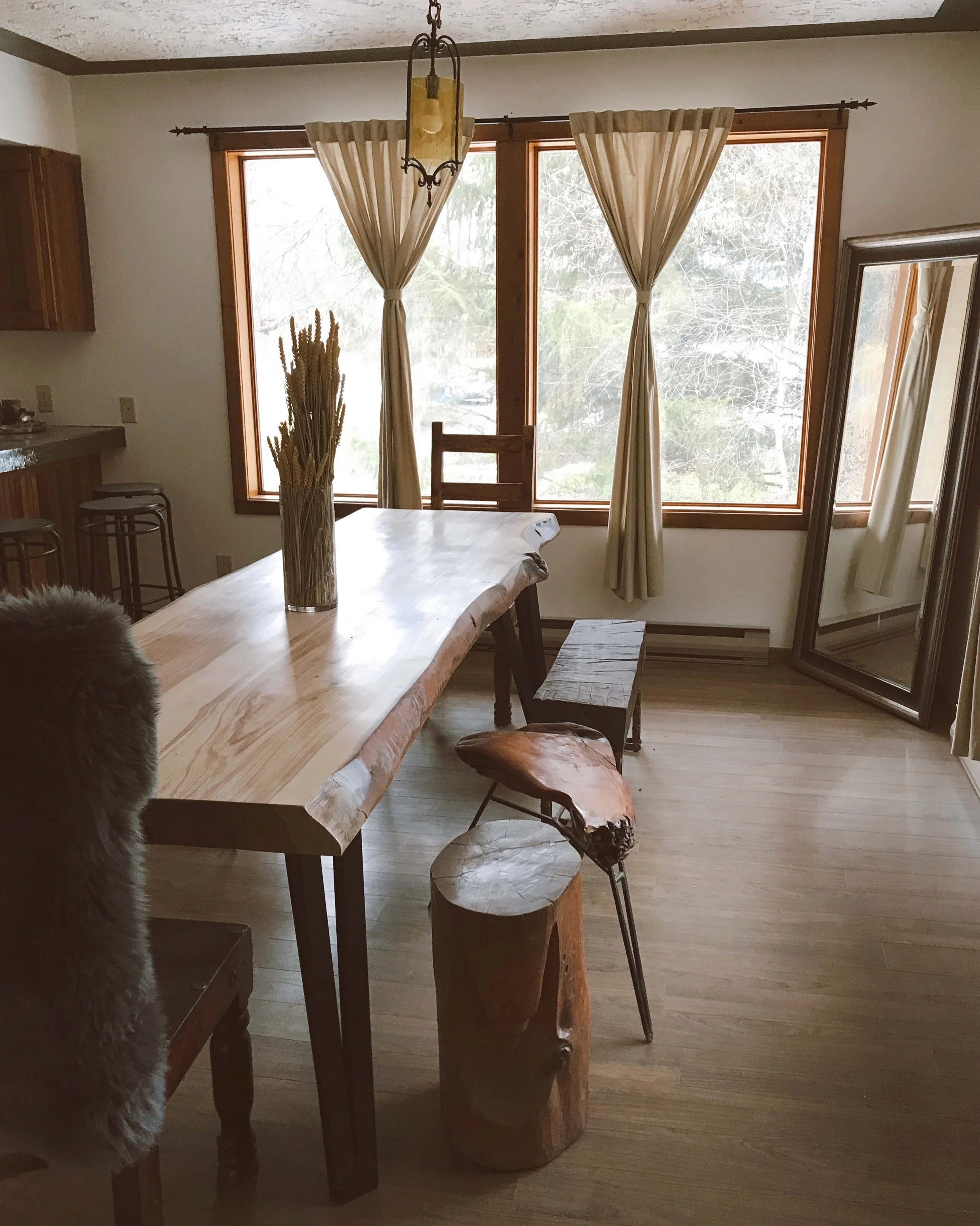 Wooden dining table with a decorative vase of dried plants, metal stools around, large windows with beige curtains, and a mirror leaning against the wall in a room with wood flooring.