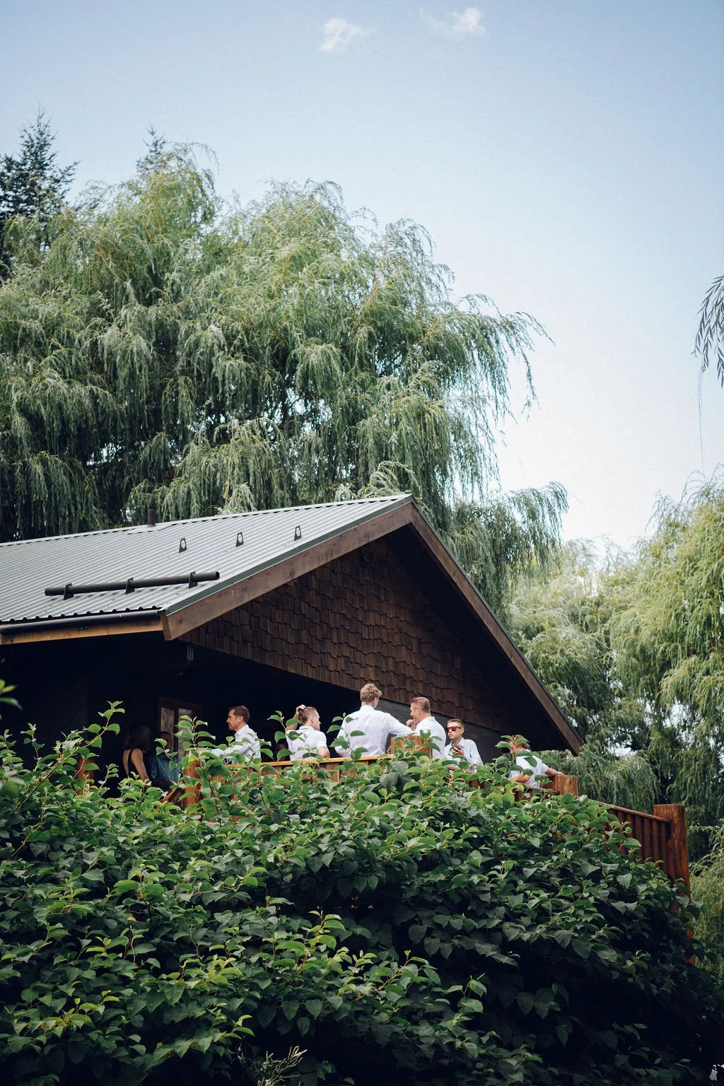 A group of people on a porch of a house surrounded by greenery, with large trees and bushes, and a metal roof on the house.