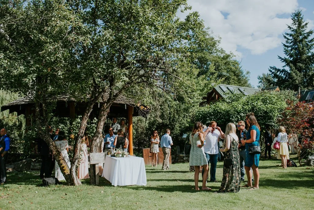People gathering outdoors in a garden with trees and a pavilion, socializing near a table with a white tablecloth.