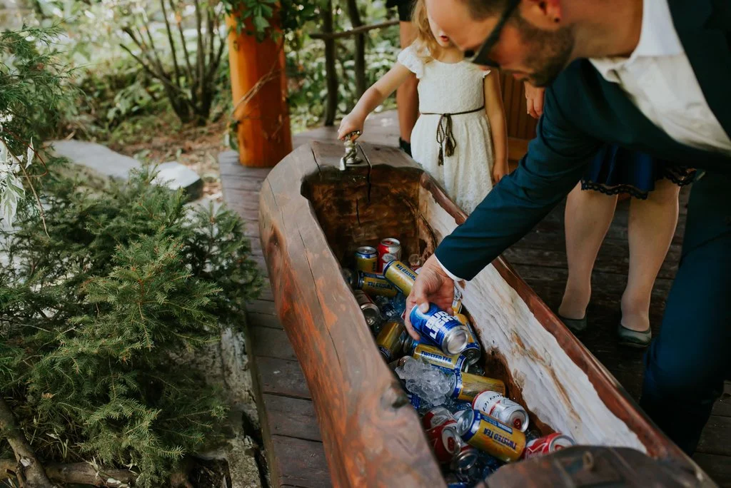 A man in a tuxedo and children are collecting empty beer and soda cans from a wooden log in an outdoor wooded area.