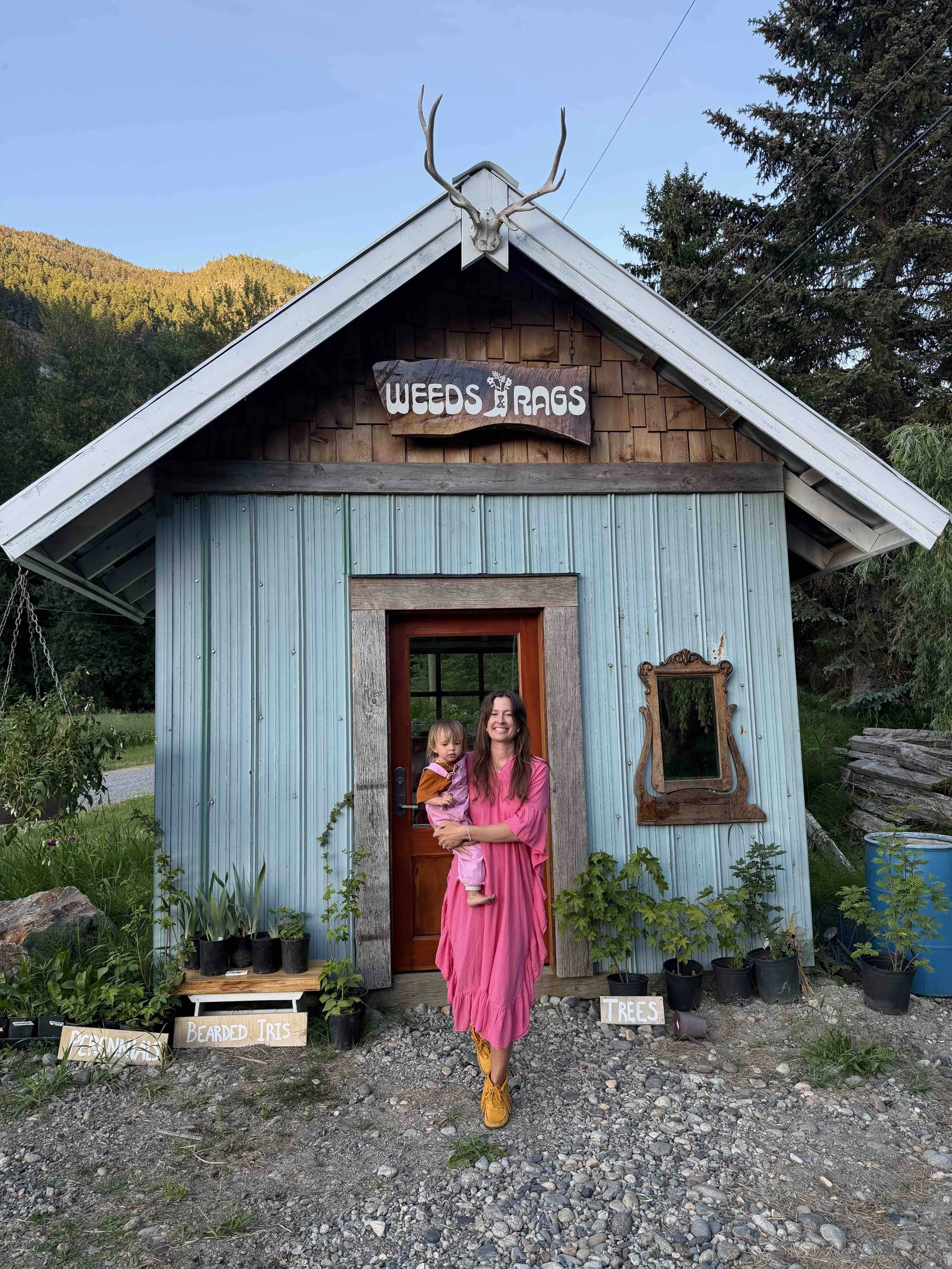 A woman in a pink dress holding a young girl in front of a small rustic building with a sign that reads 'Weeds & Rags.' Several potted plants are lined up at the base of the building, and small signs label different types of plants, such as 'Bearded Iris' and 'Trees.' A deer skull with antlers decorates the top of the building. The setting is outdoors with trees and mountains in the background.