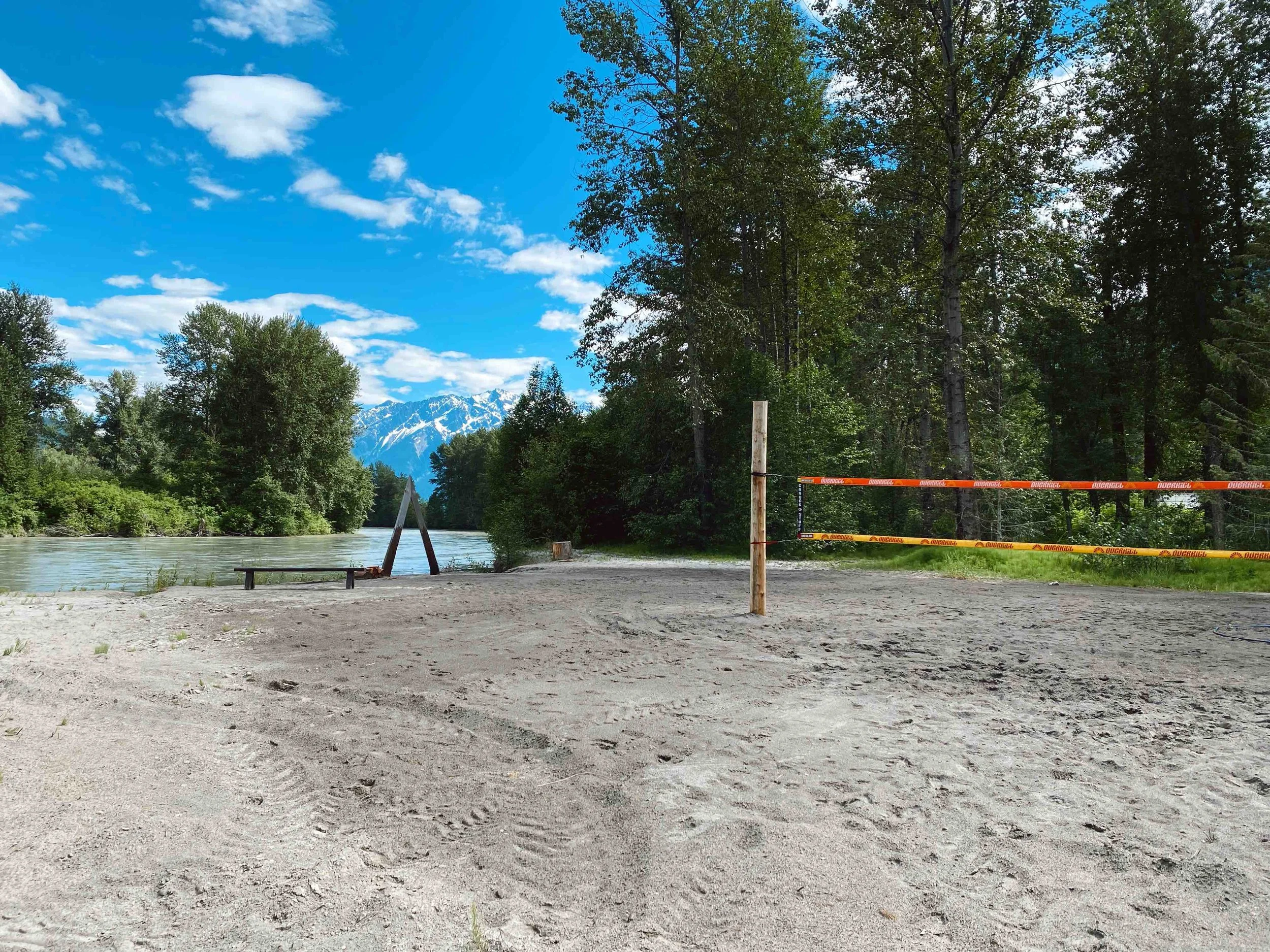 A sandy riverbank with a volleyball net set up, surrounded by tall trees, with snow-capped mountains in the distance under a blue sky with some clouds.