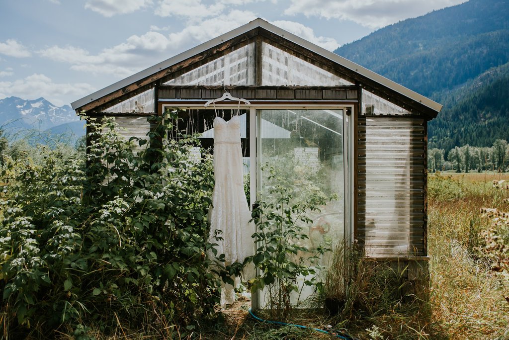 A white wedding dress hanging outside a rustic shed with a mountainous landscape in the background.
