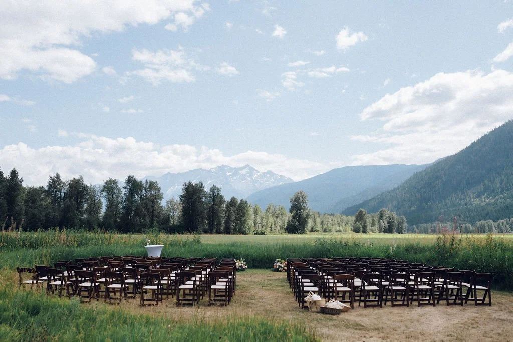 Outdoor wedding setup with rows of chairs facing a scenic mountain landscape, some flowers, and a blue sky with clouds.