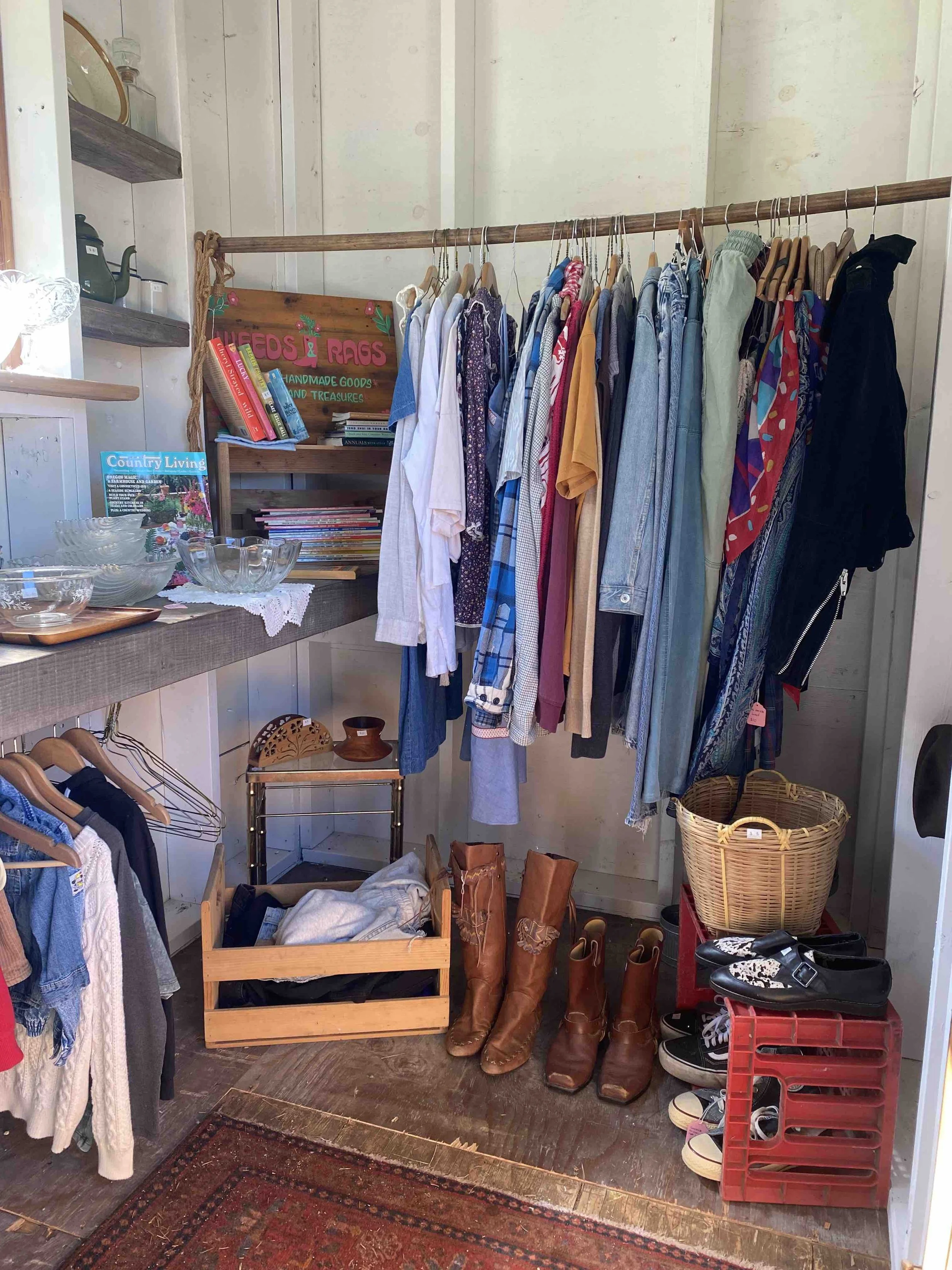 Secondhand clothing and shoes displayed in a small retail space with a clothing rack, boots, and shoes on the floor and shelves.