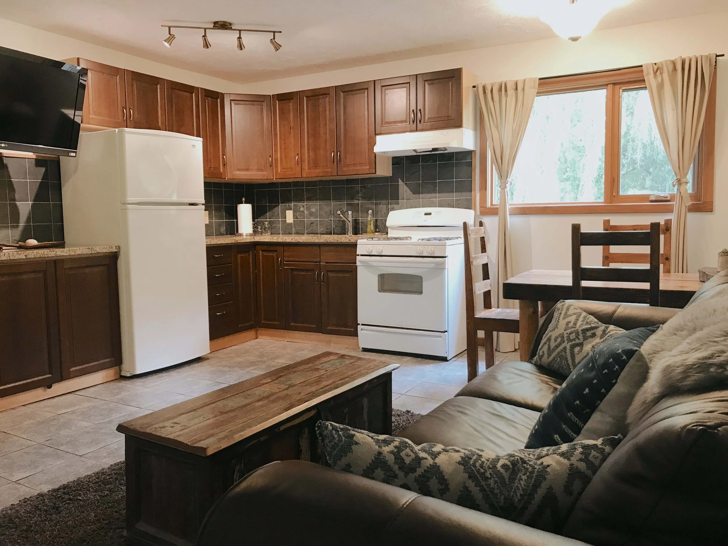 View of a cozy living room with a kitchen area in the background. Kitchen has wooden cabinets, black tiled backsplash, white stove, and a refrigerator. Large window with beige curtains allows natural light. Furniture includes a dark leather sofa with patterned cushions and a wooden coffee table. Floor is tiled, and there is a ceiling light fixture.