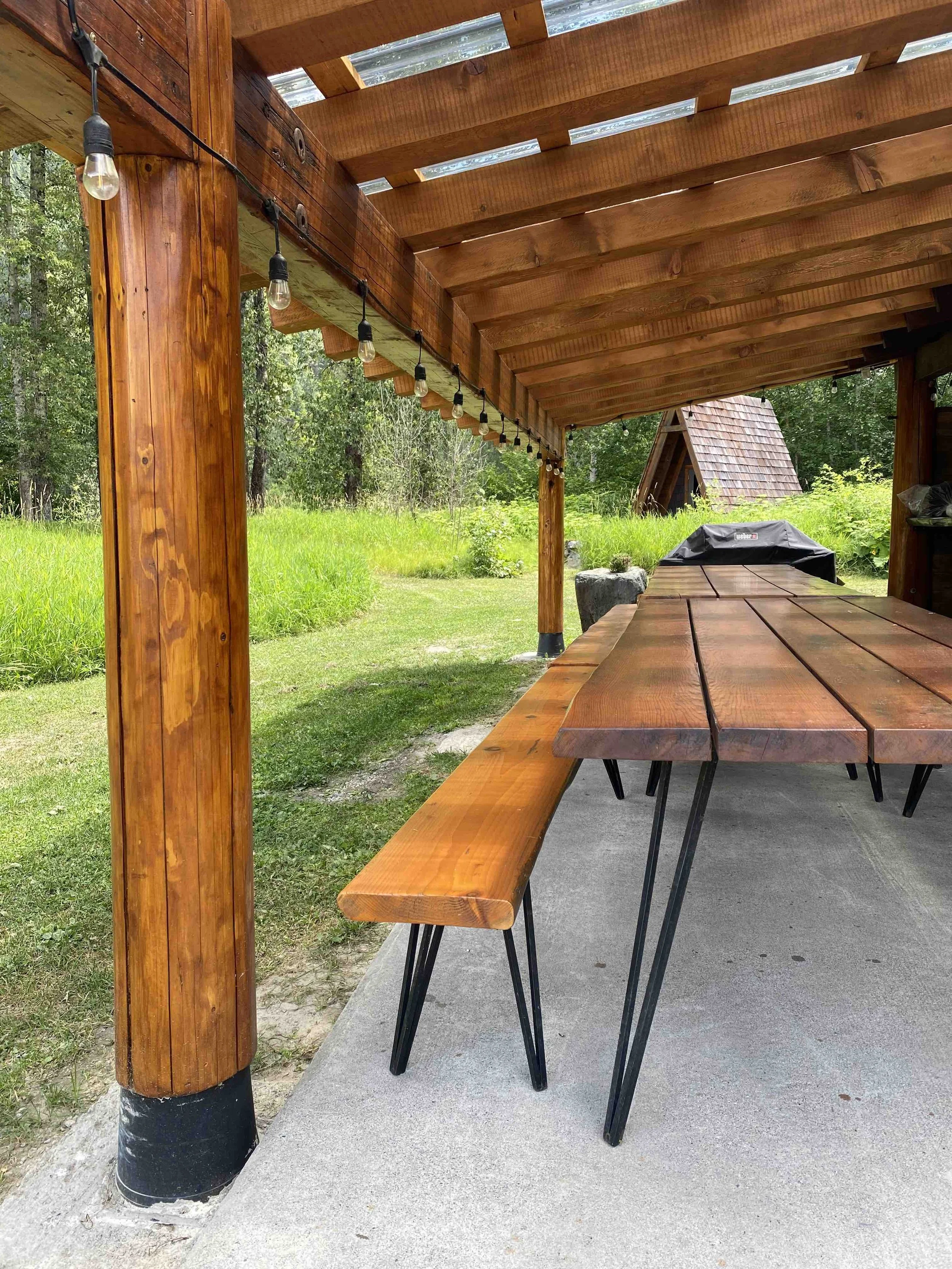 Wooden outdoor patio with a table and benches, string lights, and a grassy area with trees in the background.