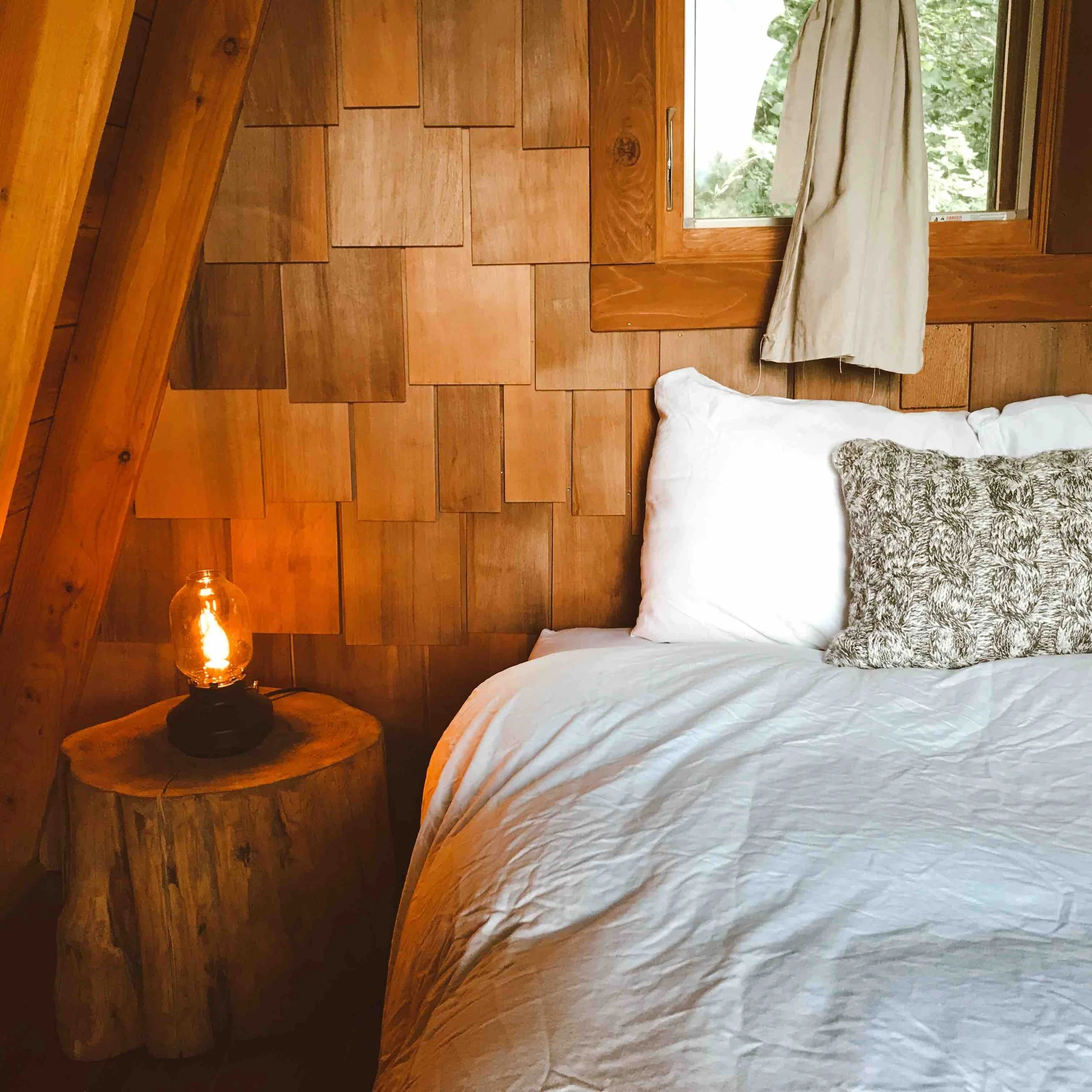 Close-up of a bedroom with wooden paneled wall, a bed with white bedding and decorative pillows, a window with beige curtains, and a bedside table with a lamp.