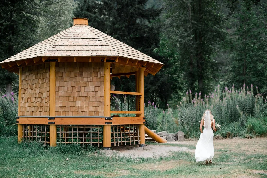 A woman in a white dress walking towards a wooden playhouse with a slide in a grassy garden with trees and tall purple flowers.