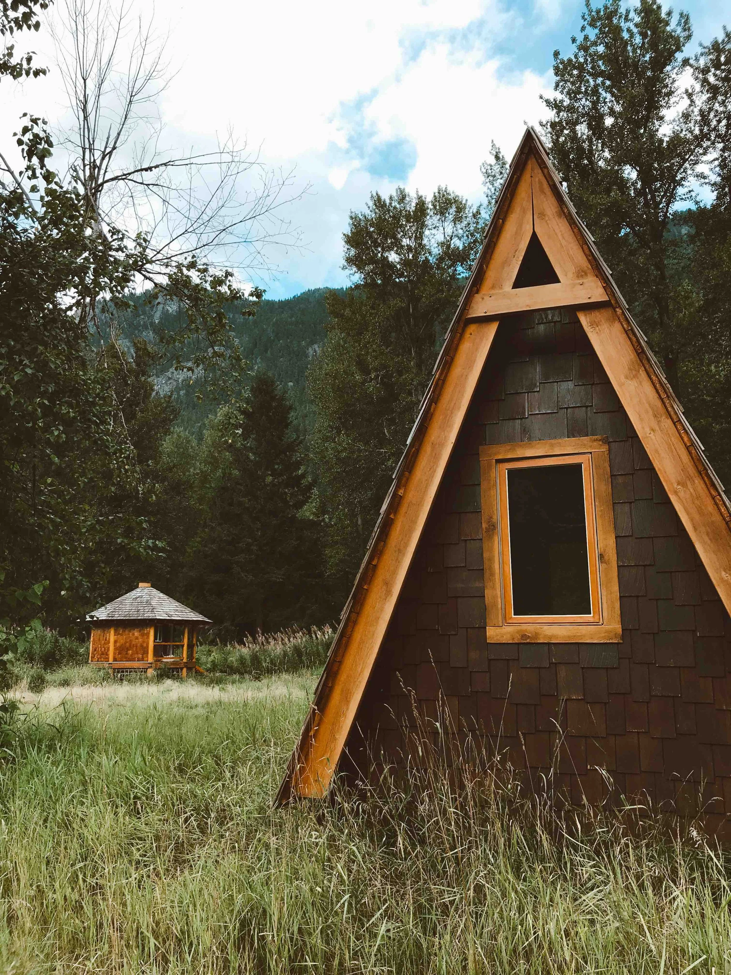 A wooden A-frame cabin with a small window, situated in a grassy clearing surrounded by trees and mountains, with a partly cloudy sky overhead.
