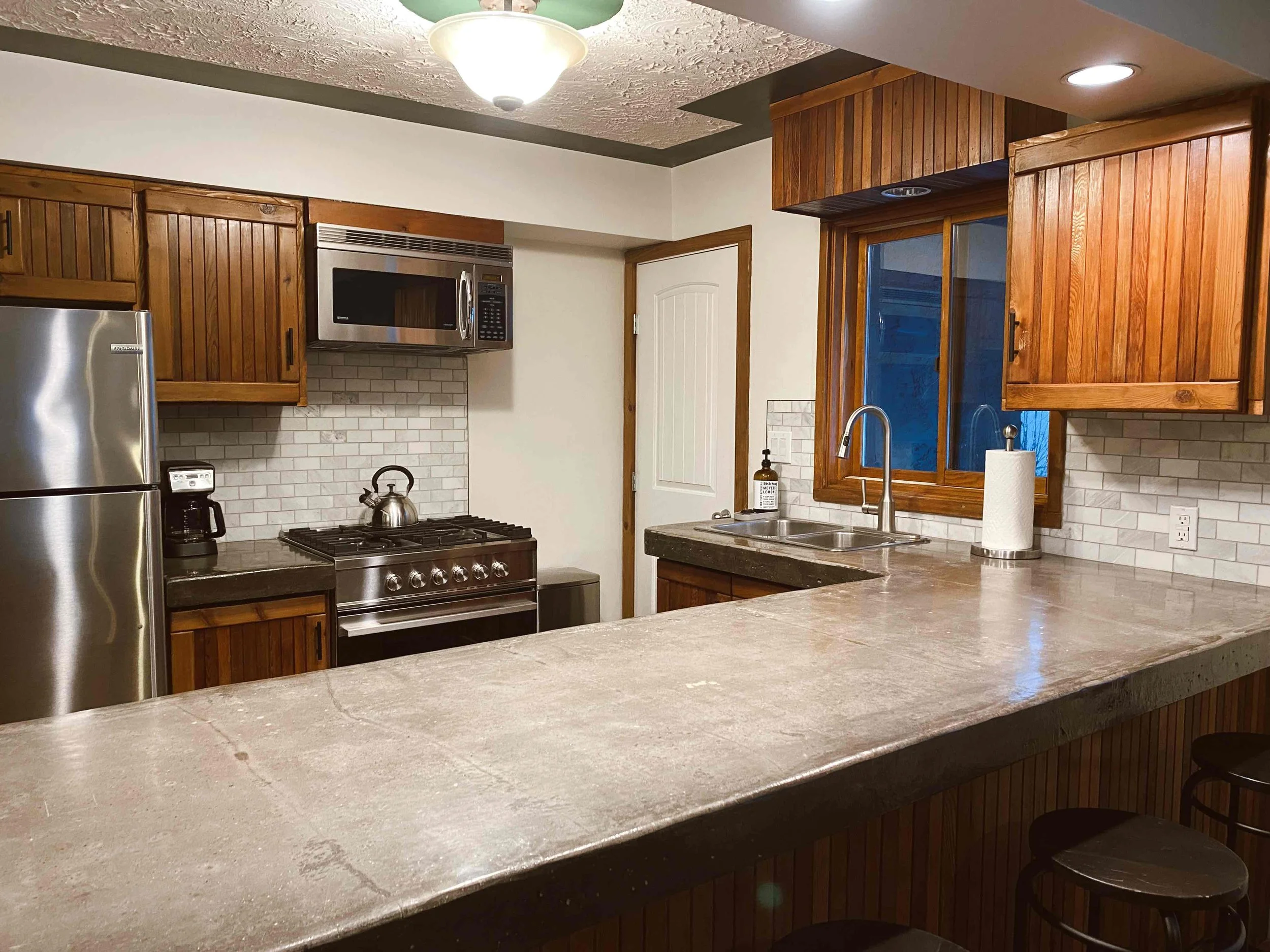 A kitchen with wooden cabinets, a granite countertop, stainless steel appliances including a fridge and microwave, a white subway tile backsplash, and a double sink beneath a window, with a paper towel roll and soap dispenser on the counter.