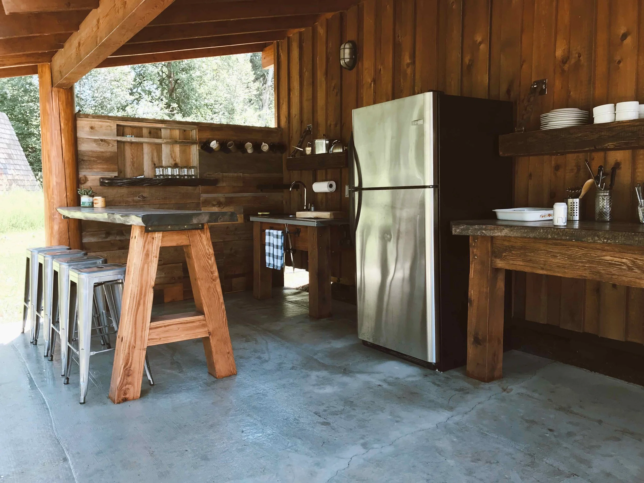 Interior of a rustic wooden kitchen with a commercial refrigerator, a wooden counter with dishes, a paper towel holder, and a window showing trees outside.