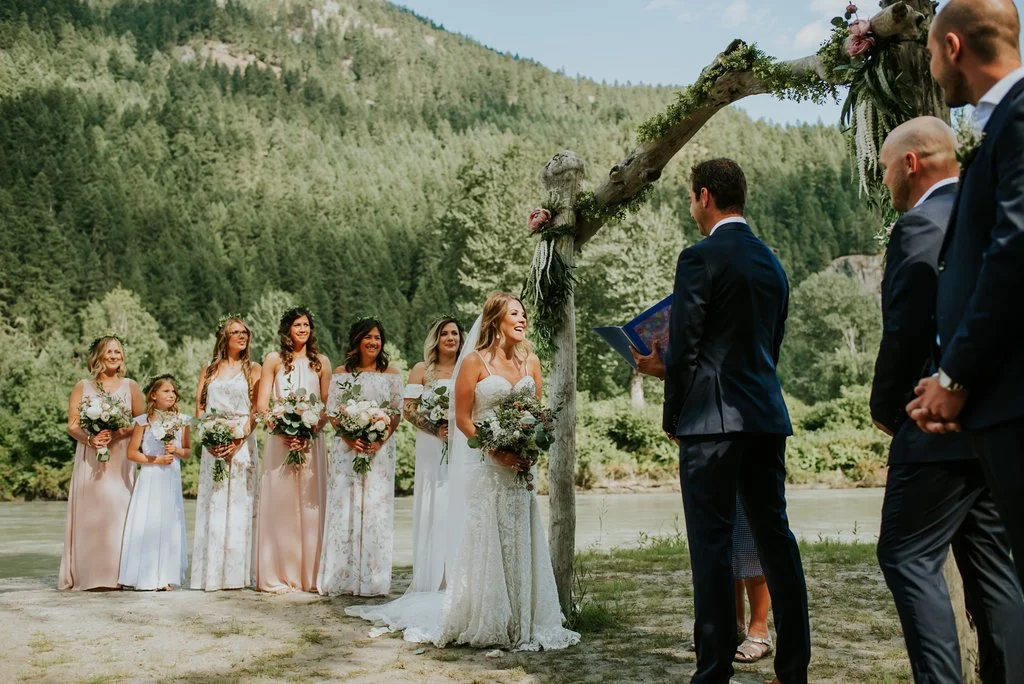 Bride and groom exchanging vows outdoors near a river, with bridesmaids and groomsmen standing nearby under a wooden arch decorated with flowers, surrounded by lush green forest.