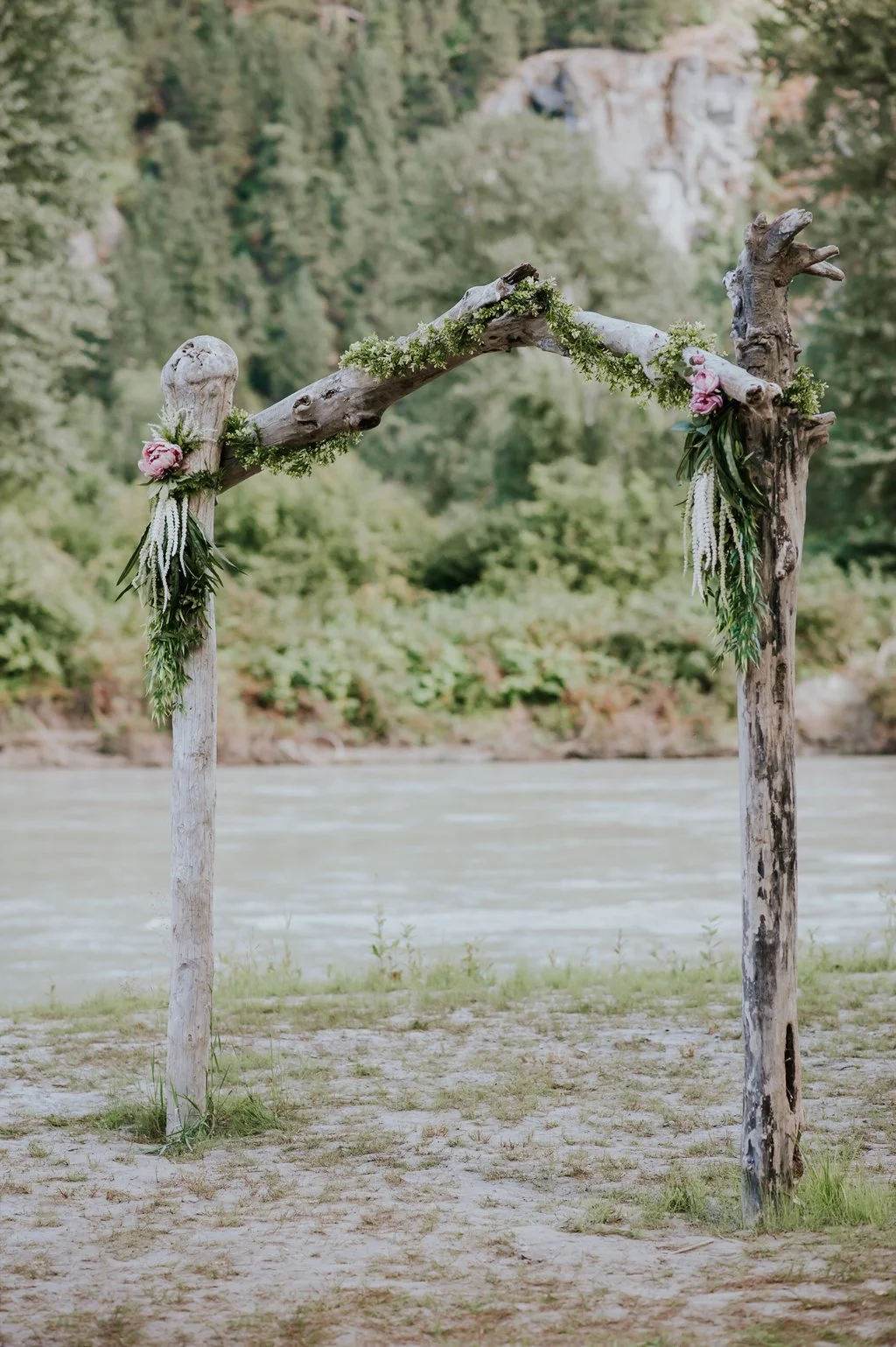 Rustic wooden wedding arch decorated with greenery and pink flowers, set outdoors near a river with a forested backdrop.