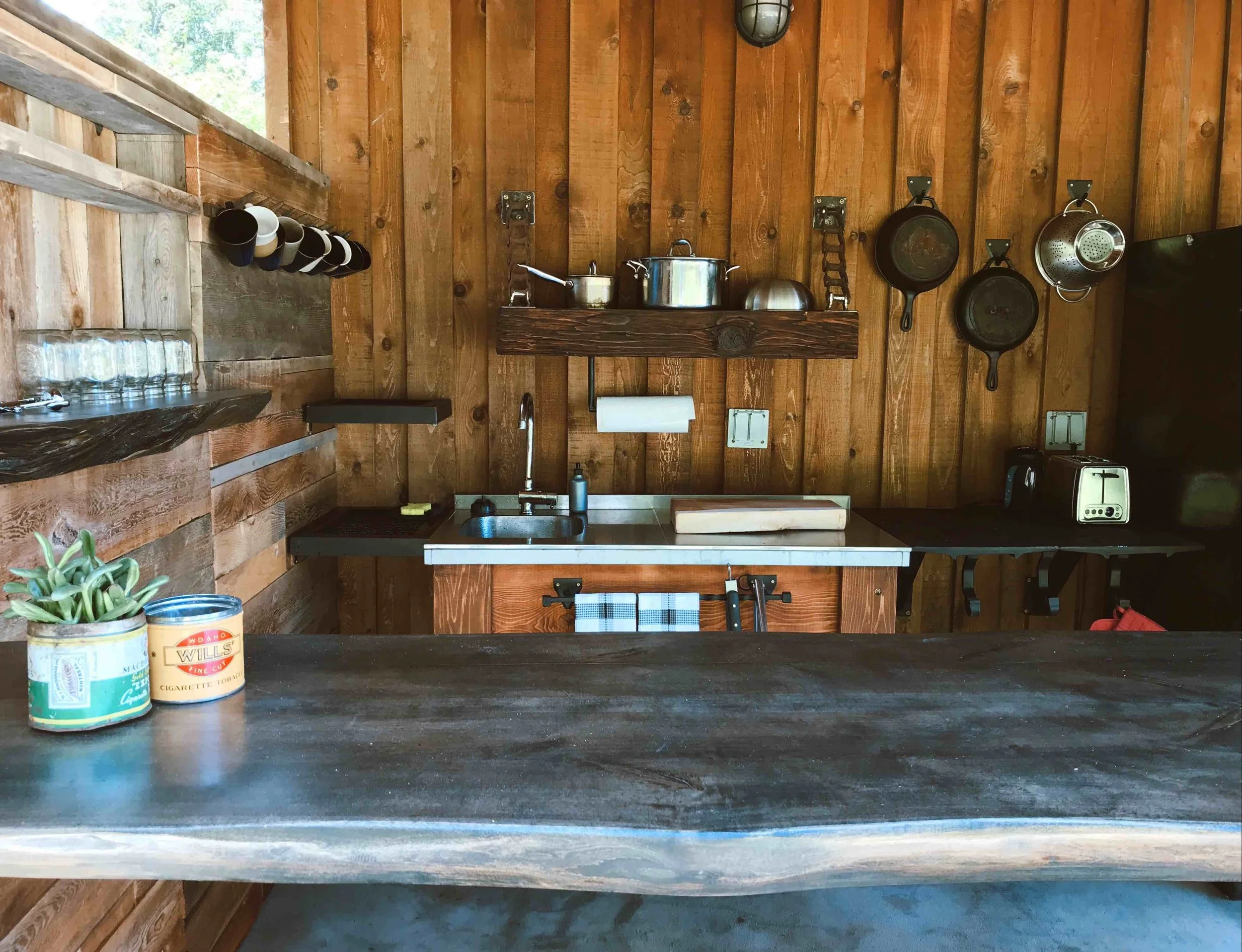 A rustic kitchen with wooden walls and a countertop. On the counter, there are two cans, one with a succulent plant and the other labeled 'Will's'. The background features hanging cast iron skillets, a chain, a small pot, and a mounted light fixture, along with a sink, paper towel holder, and a toaster.