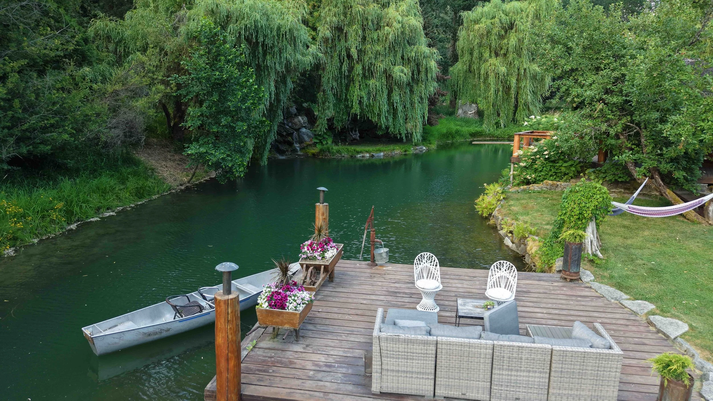 A backyard deck overlooking a pond with lush green trees and hanging willow branches. The deck has outdoor furniture, potted plants, and a small boat tied to the dock. There is a hammock on the right side among the greenery.