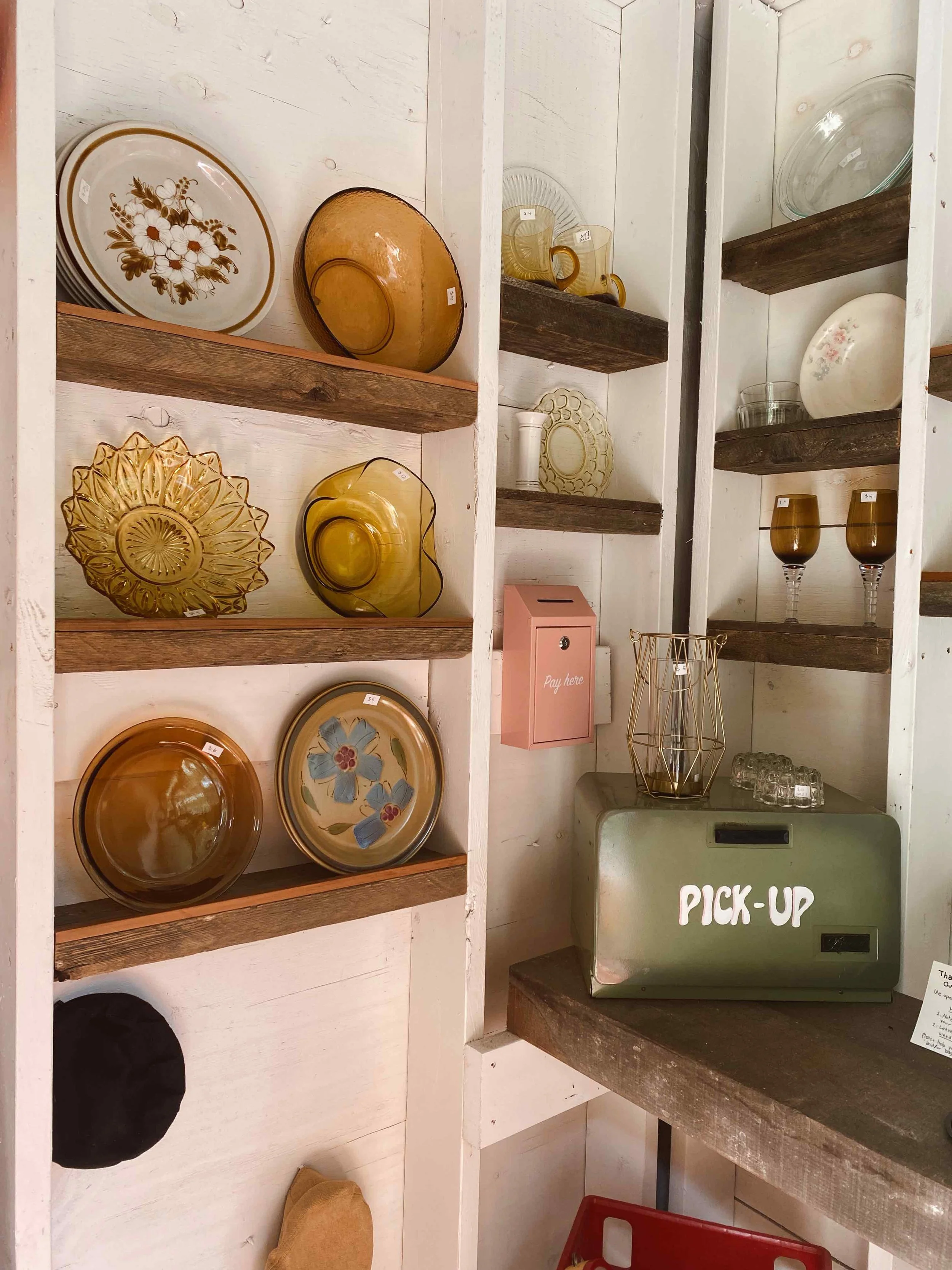 Display shelves with vintage glassware including plates, cups, and bowls, and a color-coded pick-up container labeled 'PICK-UP' in a retail store.