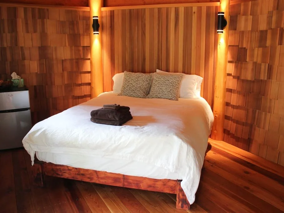 A cozy bedroom with a large bed featuring white bedding, two decorative pillows, and neatly folded towels on top. The room has wooden walls, a wooden floor, and modern wall-mounted lights.