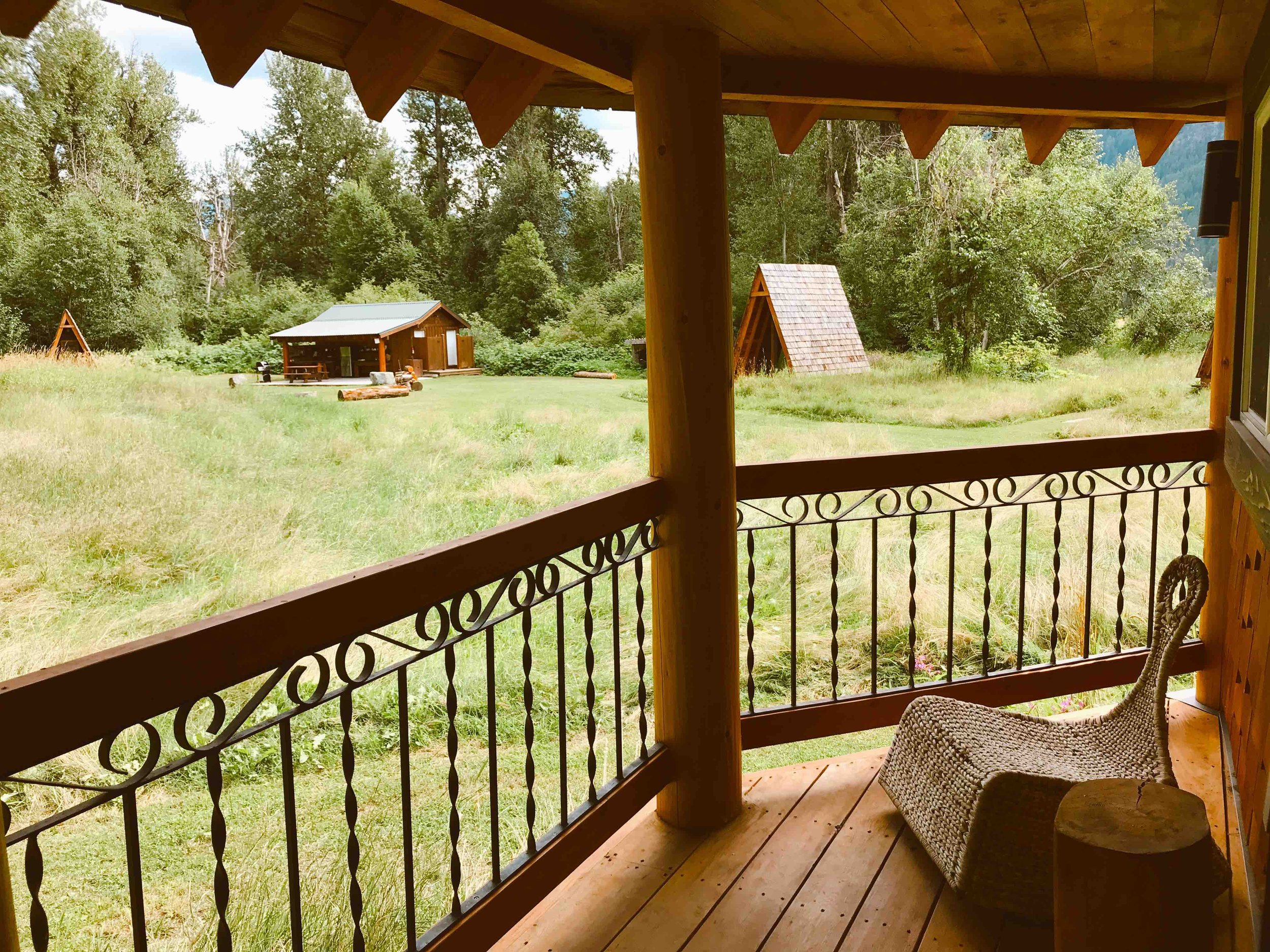 View from a porch overlooking a grassy yard with trees and small wooden structures, including an A-frame and shed, under a partly cloudy sky.