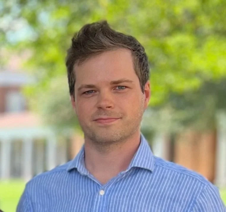 Portrait of a man with short brown hair and blue eyes, wearing a blue striped button-up shirt, standing outdoors with blurred green trees and buildings in the background.