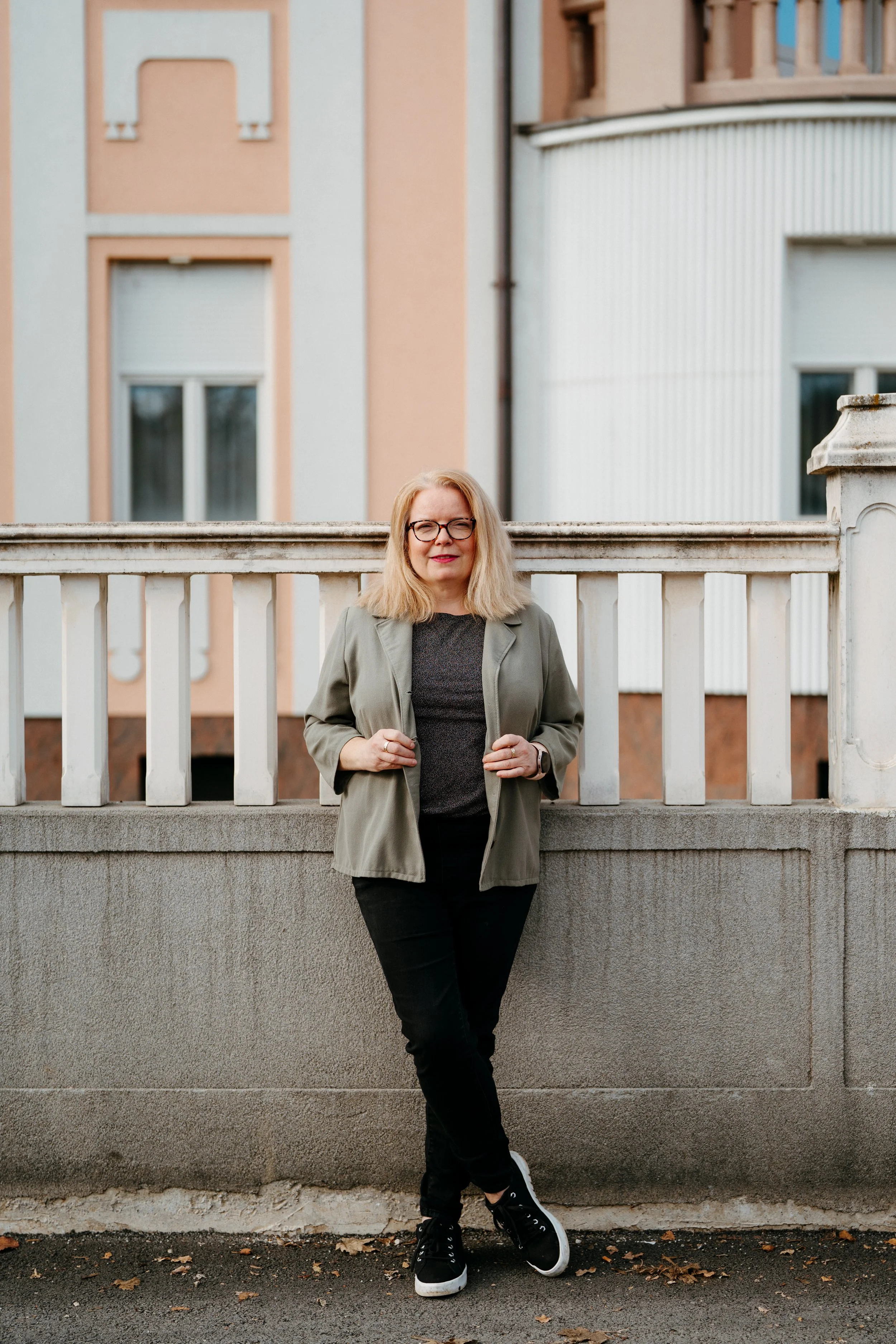 Žena plave kose i s naočalama stoji ispred dekorativnog betonskog zida u pozadini. Nosi crne hlače, zelenu jaknu, i ima neutralni  izraz lica.