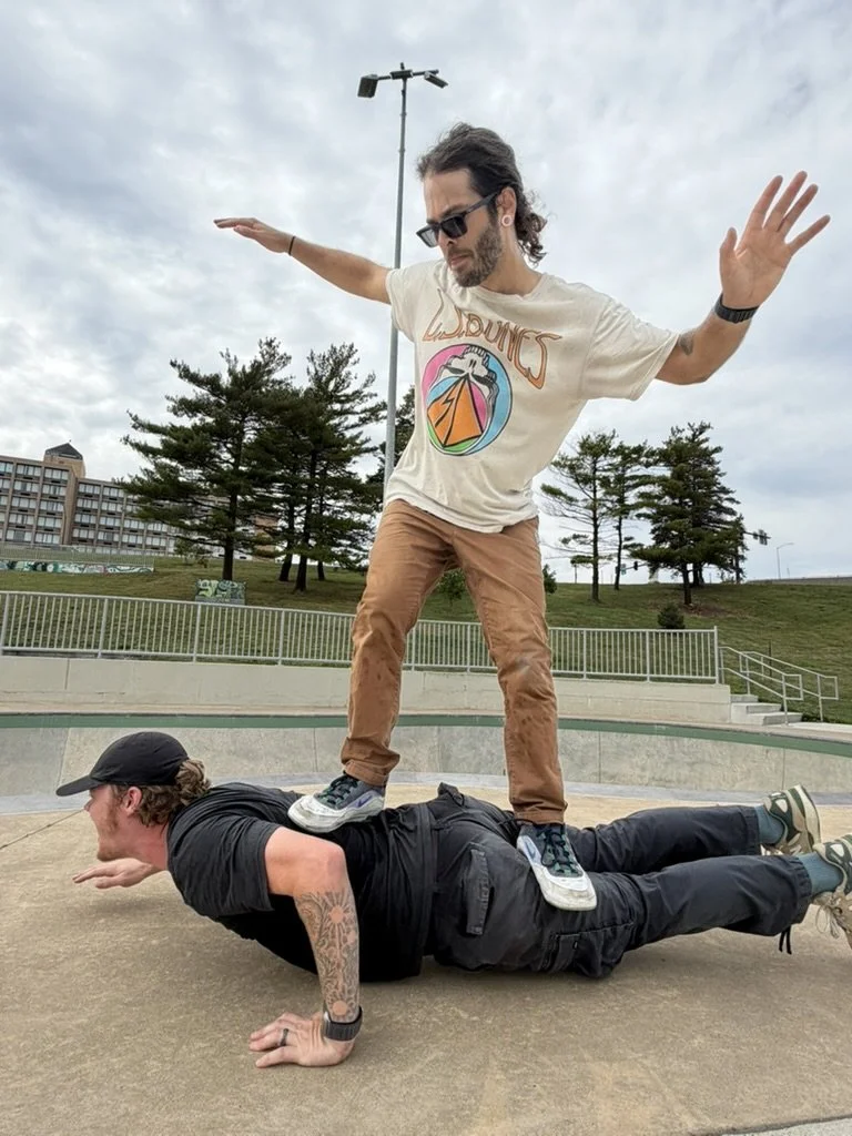 Two men performing a balancing act at an outdoor skate park. One man is lying on the ground on his stomach, supporting himself on his hands with a tattoo on his arm, while the other man stands on his back with arms extended to the sides for balance.