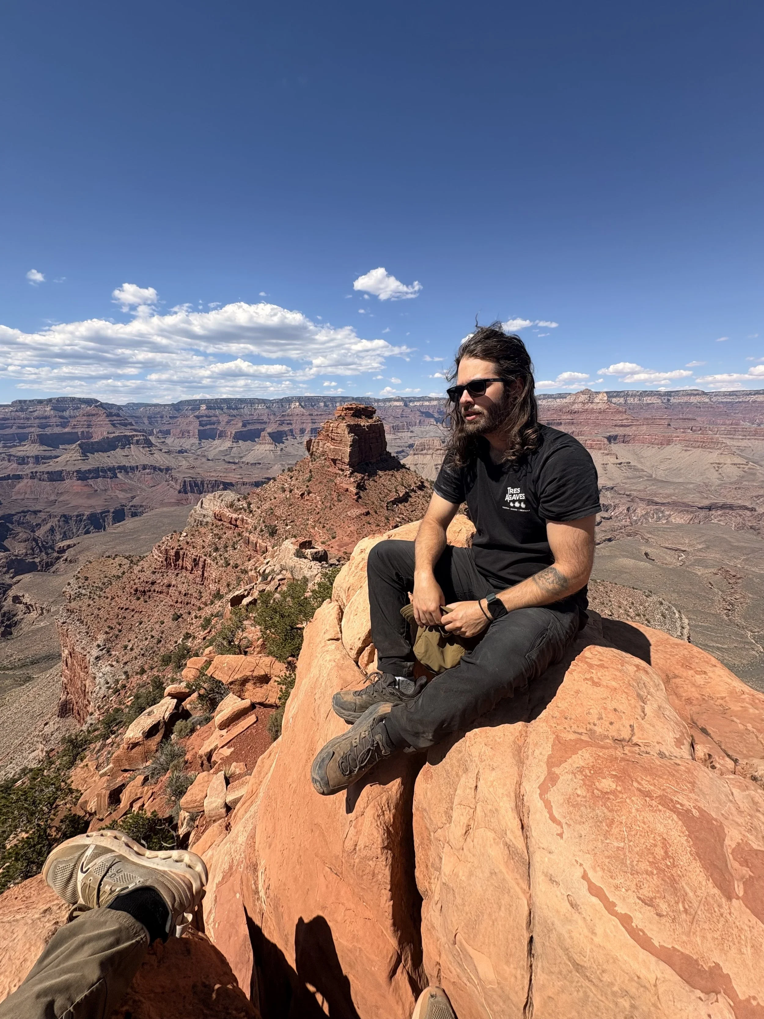 A man with long dark hair, sunglasses, black T-shirt, and outdoor gear sitting on a large orange rock formation at the Grand Canyon with a scenic view of canyon cliffs and a blue sky with clouds in the background.