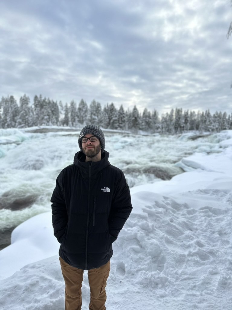 A man standing outdoors in a snowy landscape near a frozen waterfall or river, wearing a black North Face jacket, a gray beanie, and glasses, with snow-covered trees and a cloudy sky in the background.