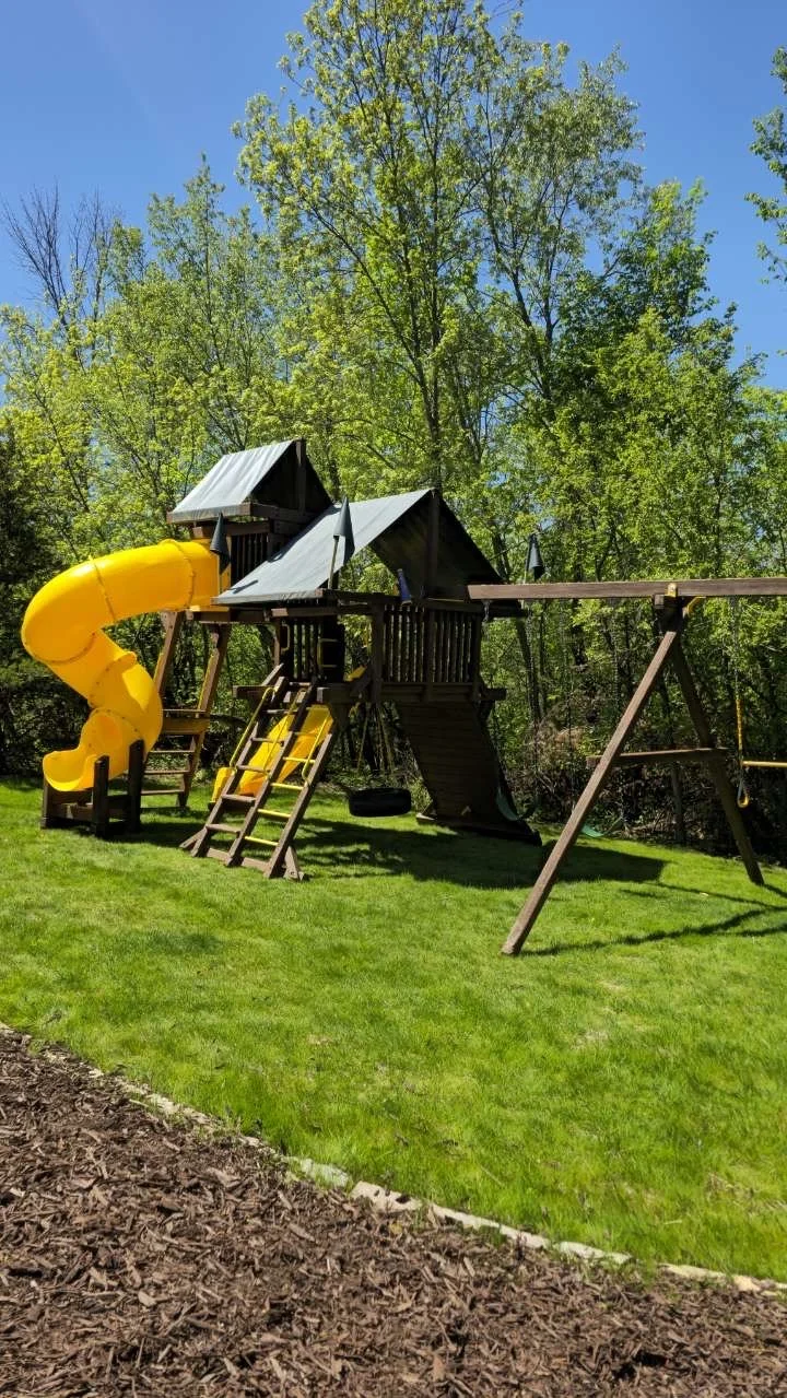A backyard playset with a yellow spiral slide, a platform, swings, and a slide, set against trees under a blue sky.