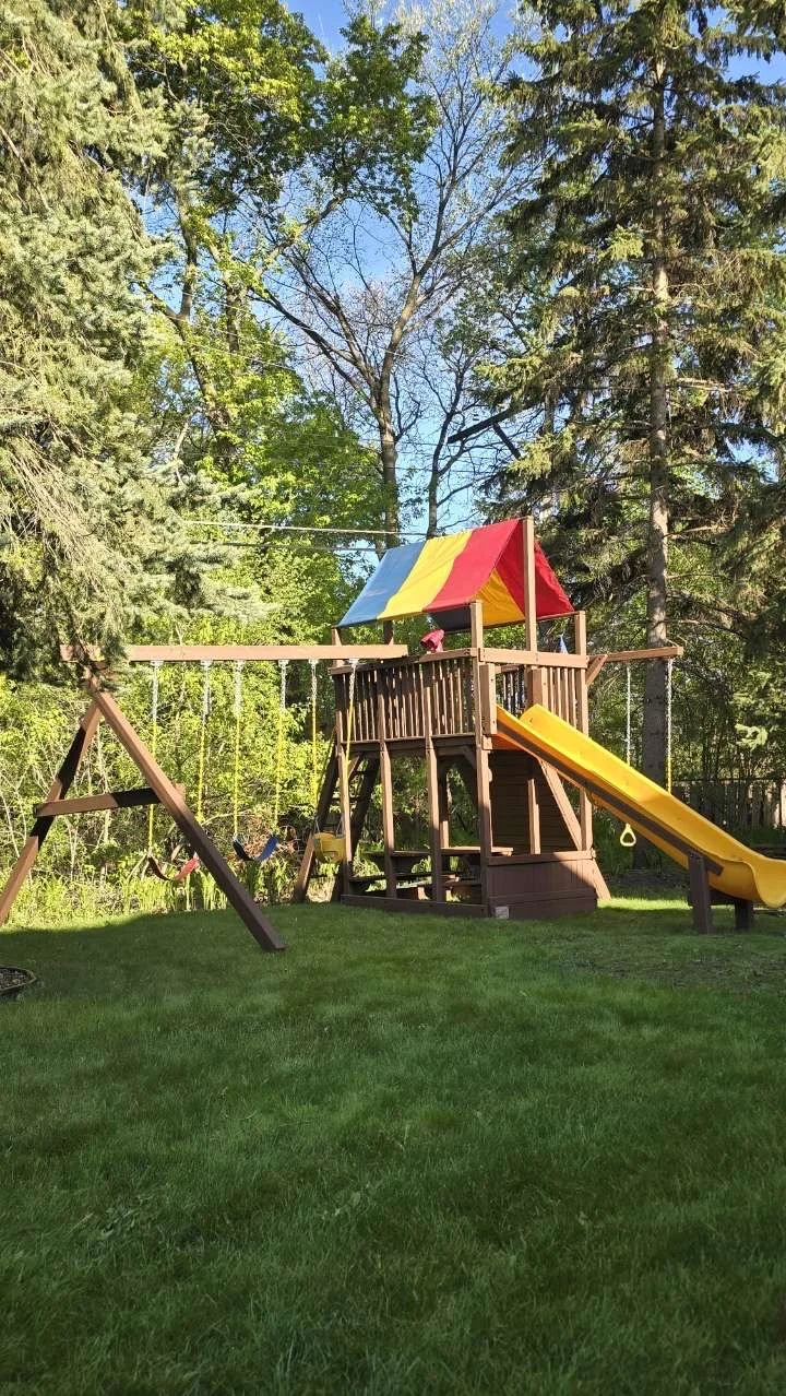 Wooden backyard playset with a slide, swings, and a colorful canopy, set on a grassy yard surrounded by trees.